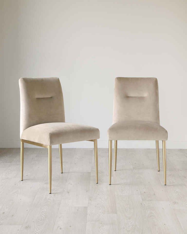 Two contemporary velvet dining chairs with a sleek design, featuring a smooth light beige upholstery and slender gold metal legs. Each chair has a high backrest with a subtle cut-out detail. The chairs are positioned on a light wooden floor against a clean white wall, highlighting their elegance and modern aesthetic.