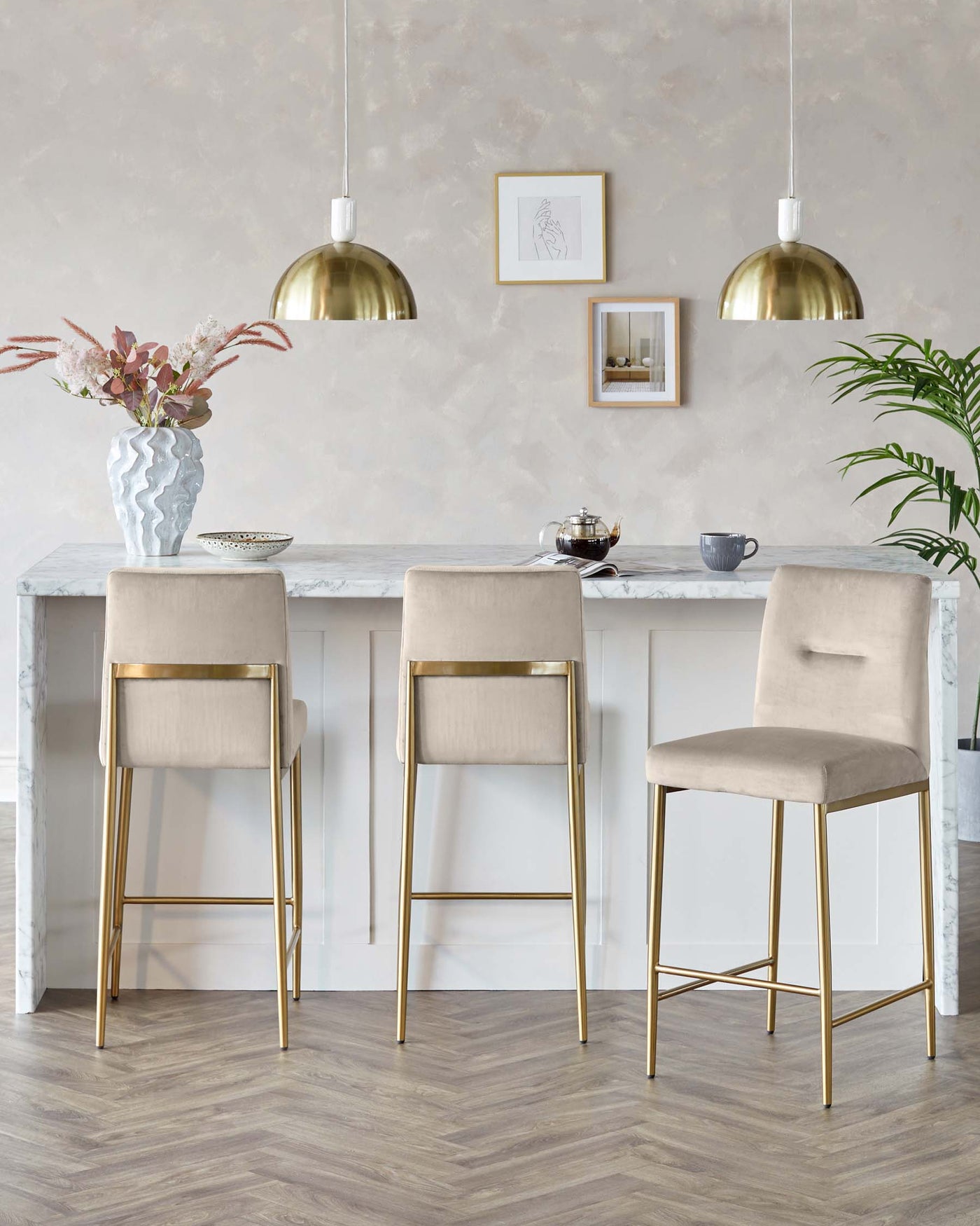 Elegant modern kitchen space featuring a white marble-top bar table with gold-finished legs and three taupe upholstered bar stools with sleek gold frames.