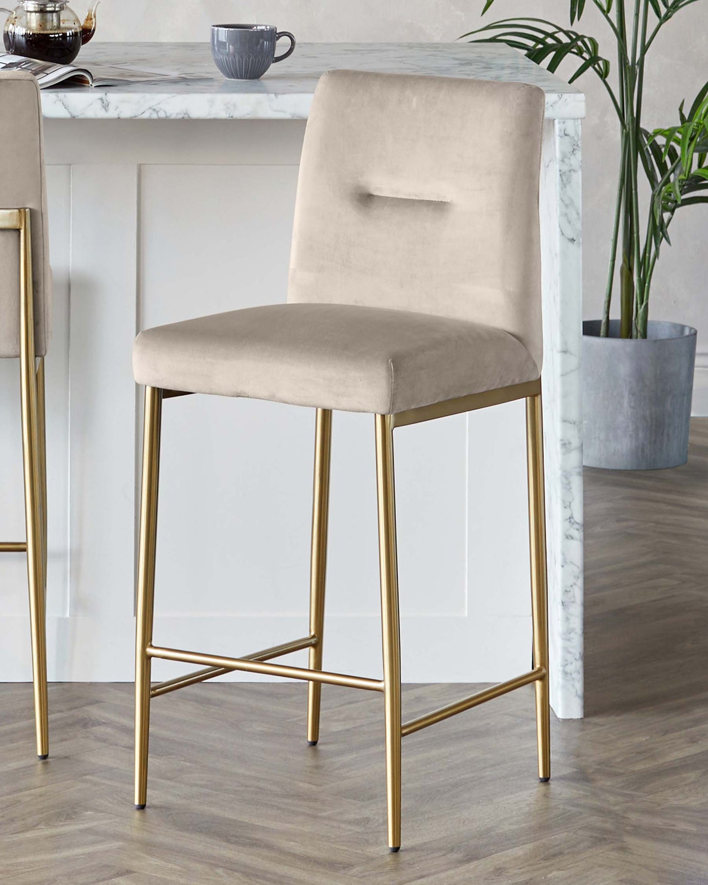 Elegant modern bar stool featuring a beige velvet upholstered seat and backrest with a convenient handle cut-out, complemented by sleek gold-finished metal legs in a geometric design.