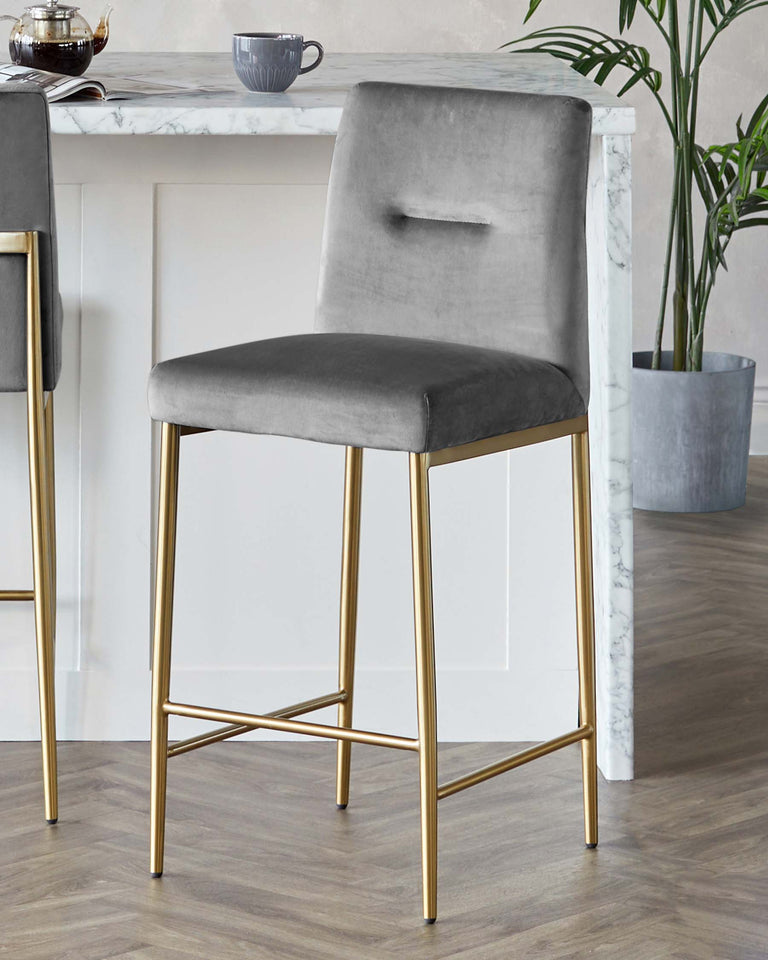 Elegant modern bar stool with a plush, grey velvet upholstered seat and backrest, supported by a sleek, gold-finished metal frame with geometric accents. The stool is positioned at a white marble top kitchen island in a contemporary setting, enhanced by natural light and complemented by similar decor.