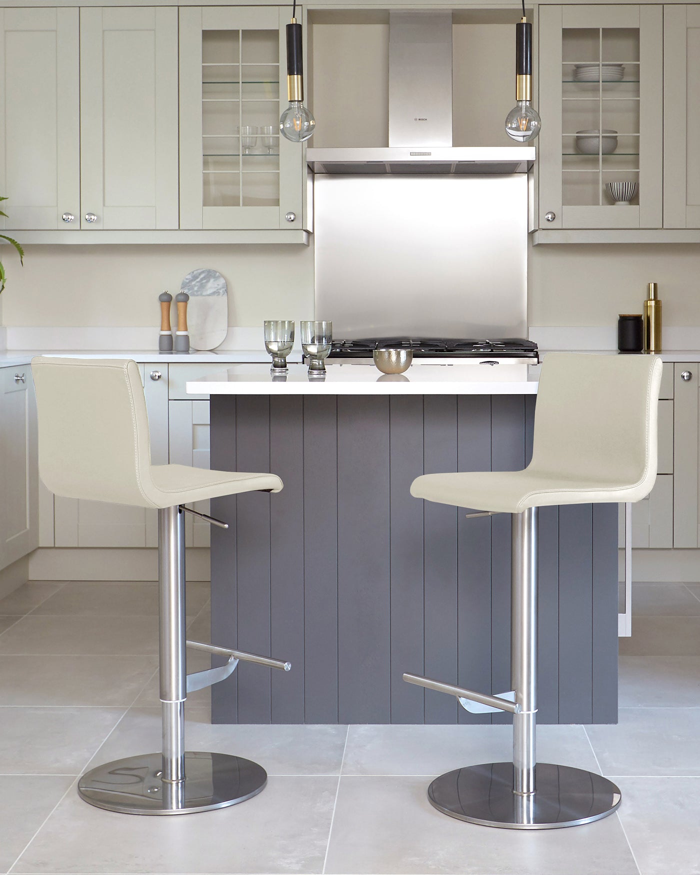 Modern kitchen with two adjustable-height bar stools featuring cream upholstery and a sleek chrome base with footrests, positioned at a kitchen island with a grey countertop.