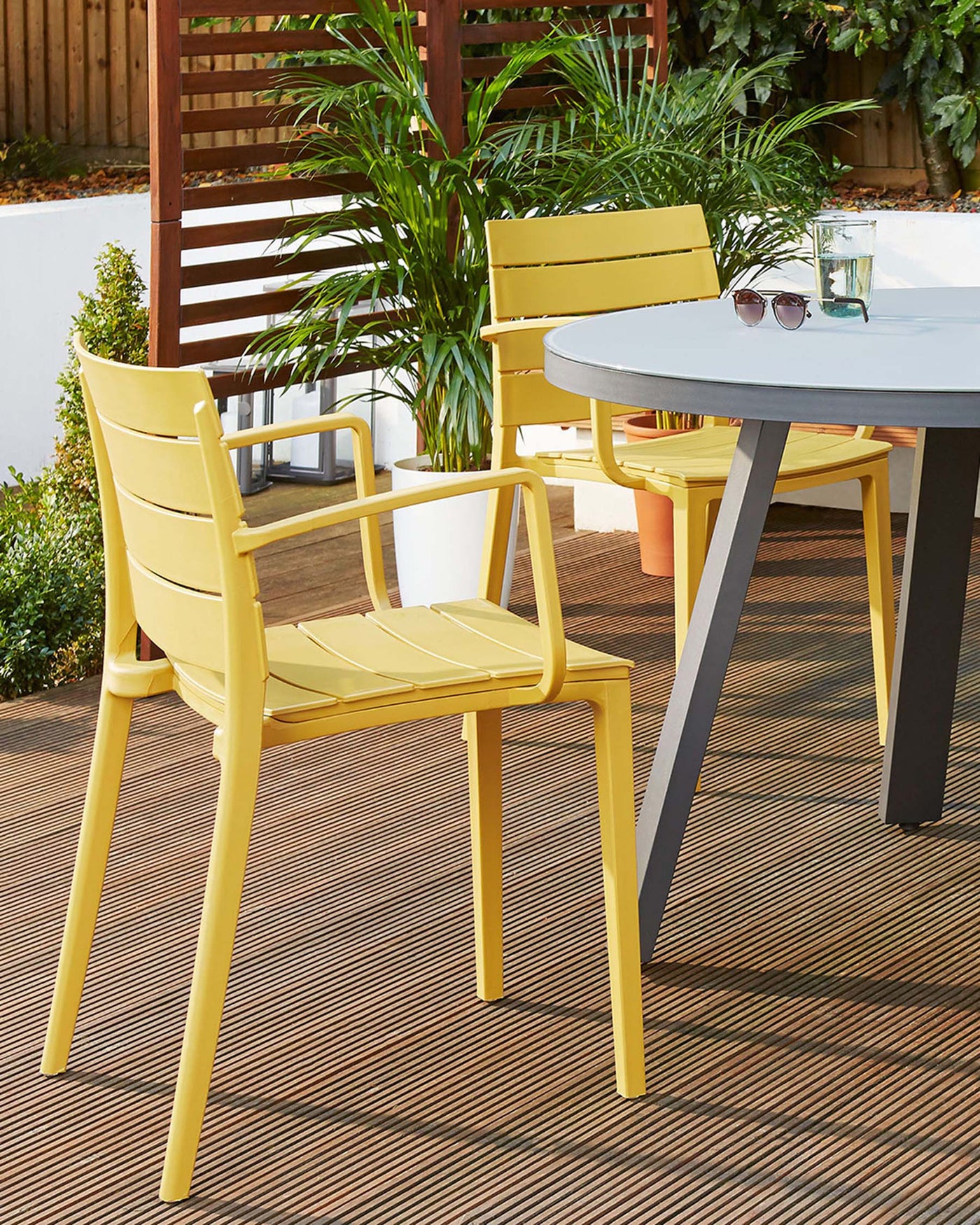Modern outdoor furniture set featuring a sleek round table with a white top and grey tapered legs, accompanied by vibrant yellow armchairs with horizontal slat backing on a wooden deck.