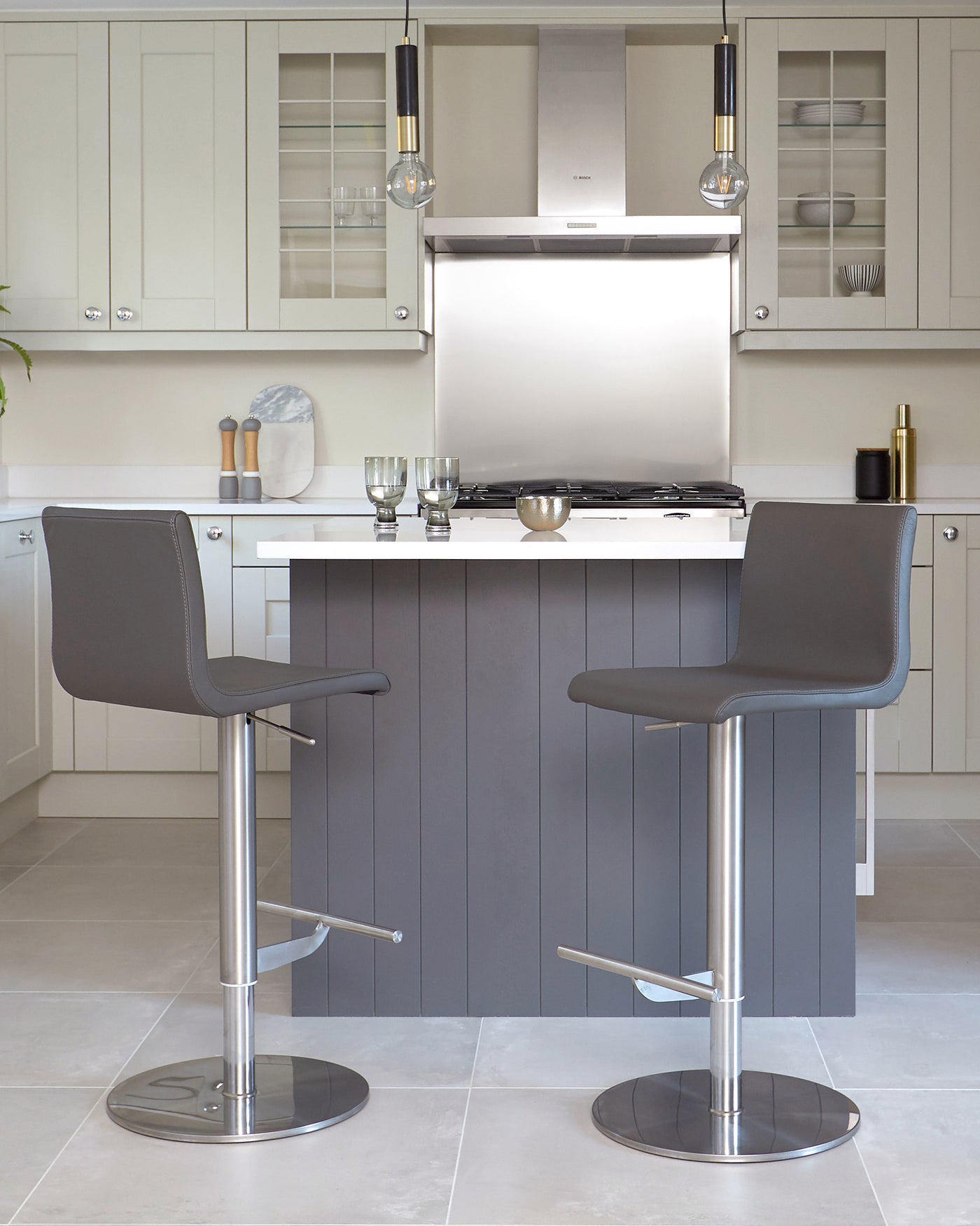 Two modern adjustable-height bar stools with footrests, featuring sleek grey upholstery and chrome bases, situated in a contemporary kitchen setting.