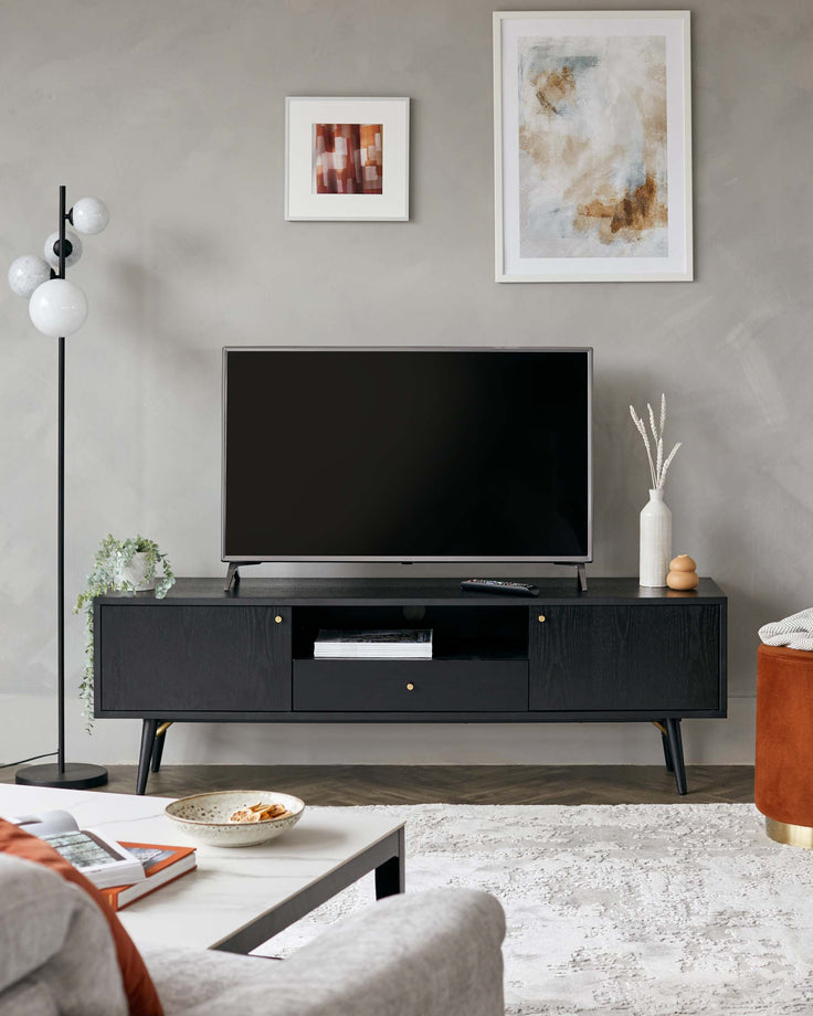A modern minimalist living room featuring a sleek black TV console with slender legs and handle-free drawers, accompanied by a complementary low-profile grey fabric sofa. The room is anchored by a light-hued area rug and accented by a simple white coffee table with metallic framing, next to a burnt orange round ottoman with a velvety texture and a golden base. The space is also defined by a tall black floor lamp with globe shades.