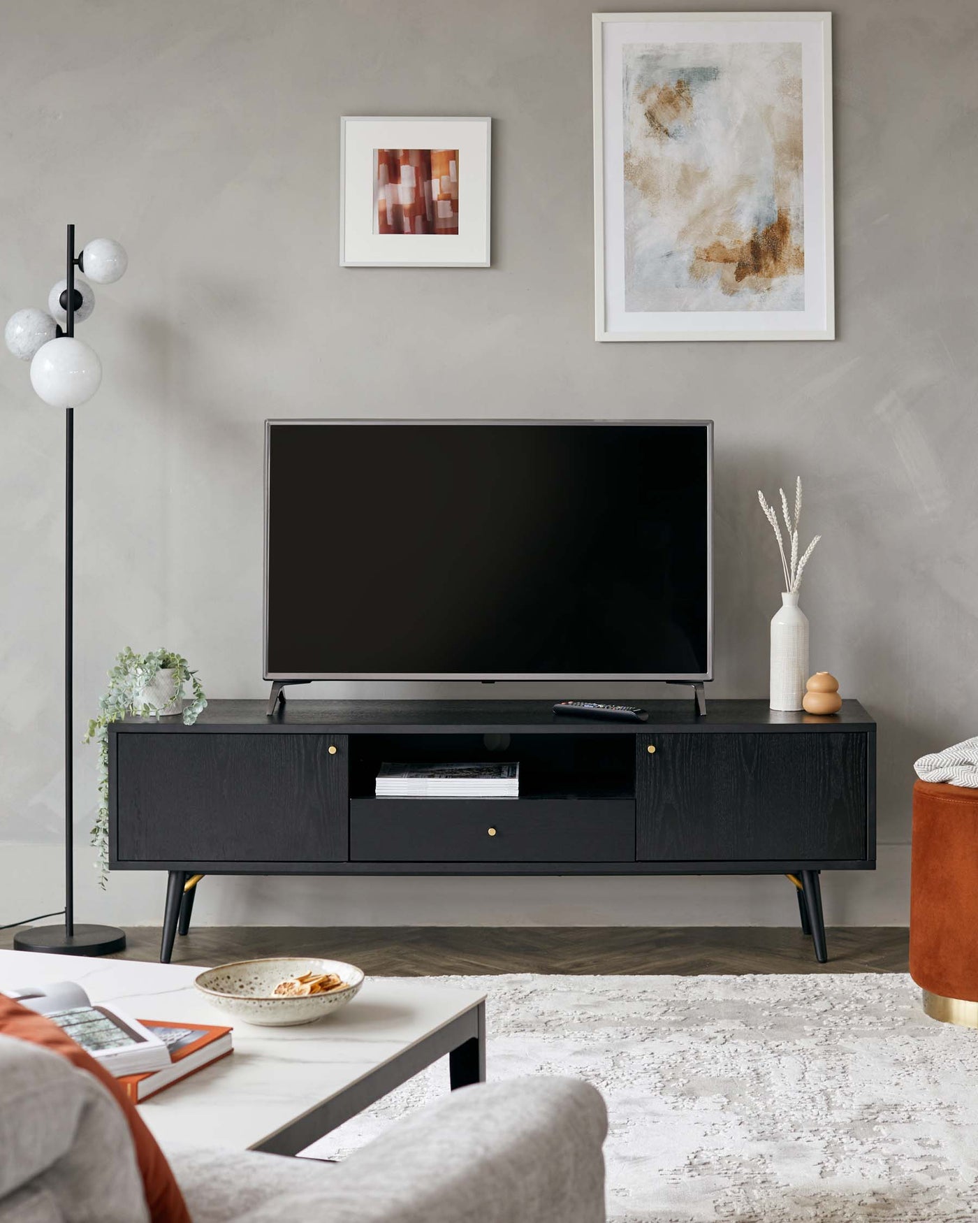 Modern minimalist living room with textured grey walls featuring a sleek black TV console with clean lines and solid rectangular shapes, standing on slender metallic legs. A low-profile round side table with velvety orange upholstery and a gold base complements the scene, and a rectangular coffee table with a grey surface and thin metal frame anchors the space, subtly blending with an off-white textured area rug.