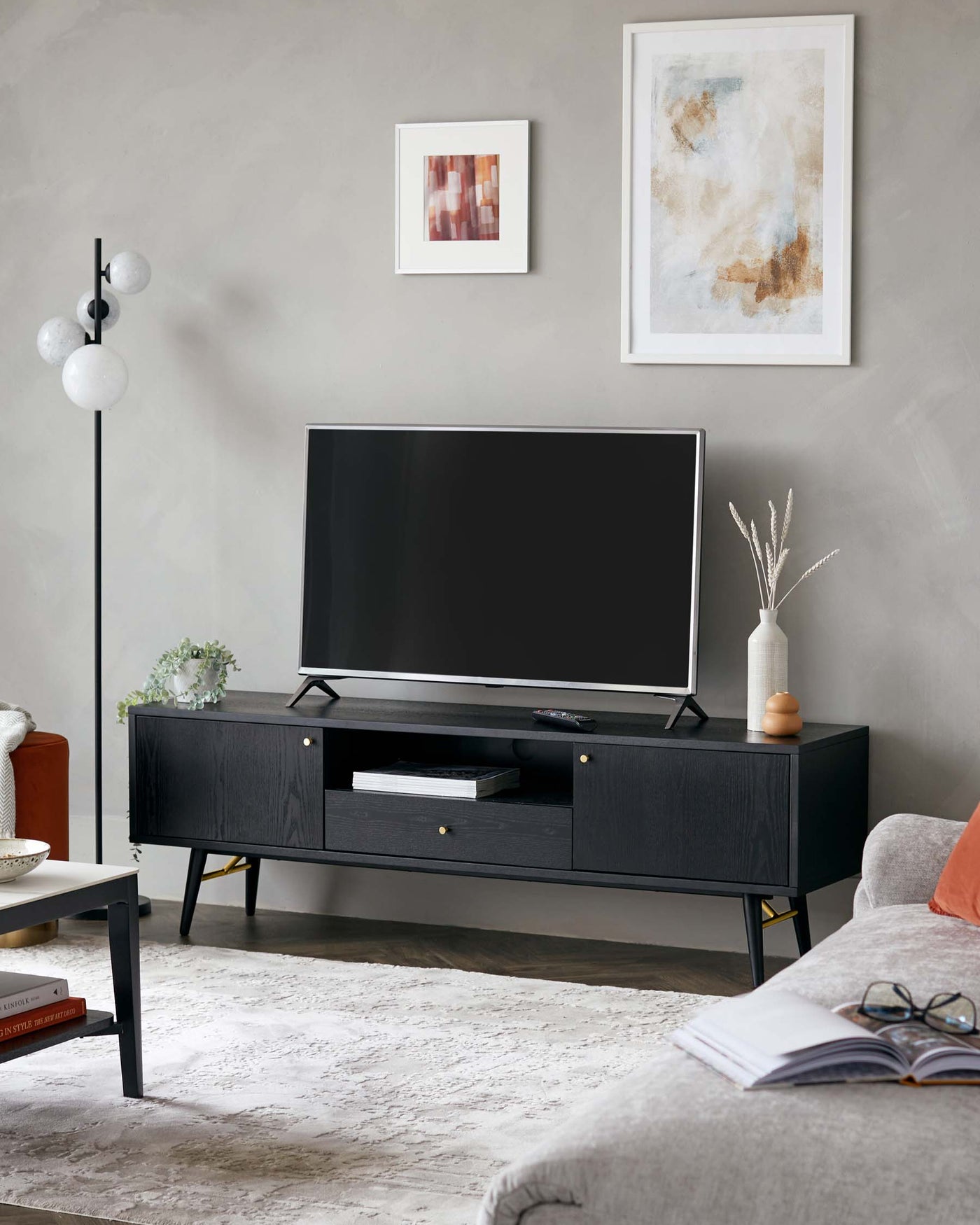 A contemporary black TV stand with clean lines, featuring open shelving and closed compartments for storage, stands on slender metal legs. In the foreground, a portion of a light grey fabric sofa with a tufted cushion is visible, accompanied by a small round side table with a white top and wood legs. The scene is completed with an accent floor lamp that has a slender pole and three spherical shades.