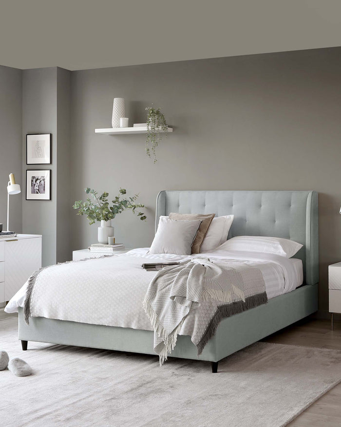 Elegant bedroom furniture set including a pastel green upholstered bed frame with a tufted headboard, white and grey bedding, a white floating shelf on the wall, a small white bedside table with a drawer, and a modern white table lamp.