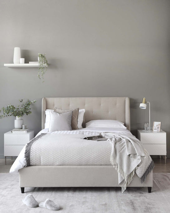 Contemporary bedroom furniture set featuring a neutral-toned upholstered bed with a tufted headboard, complemented by two white modern nightstands with geometric detailing. A minimalist white floating shelf is placed above the bed, and a sleek white lamp with a gold accent extends elegantly from one of the nightstands.