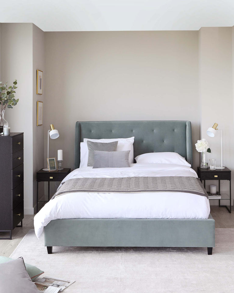 An elegant bedroom display featuring a plush, upholstered king-sized bed in a soft sage green with button tufted headboard detail. The bed is outfitted with crisp white bedding, a textural grey throw, and complementary decorative pillows. Flanking the bed are two black nightstands with a sleek, minimal design, each adorned with stylish table lamps and small decorative items. The scene exudes a serene, contemporary aesthetic.