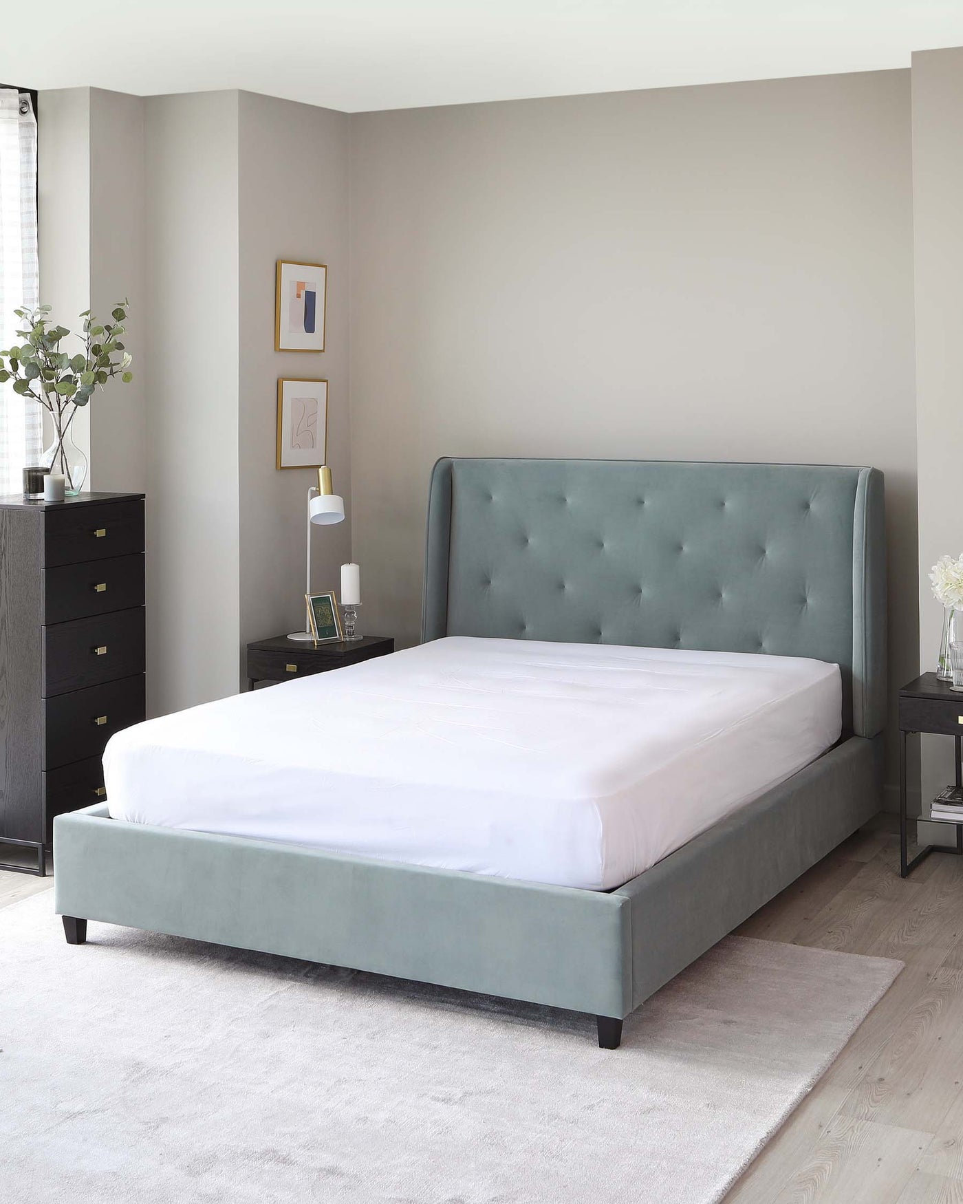 A contemporary bedroom featuring a grey upholstered bed with a tufted headboard, next to a matching grey nightstand with black accents, and a sleek black drawer dresser. The room is completed with minimalist decor and a soft grey area rug.