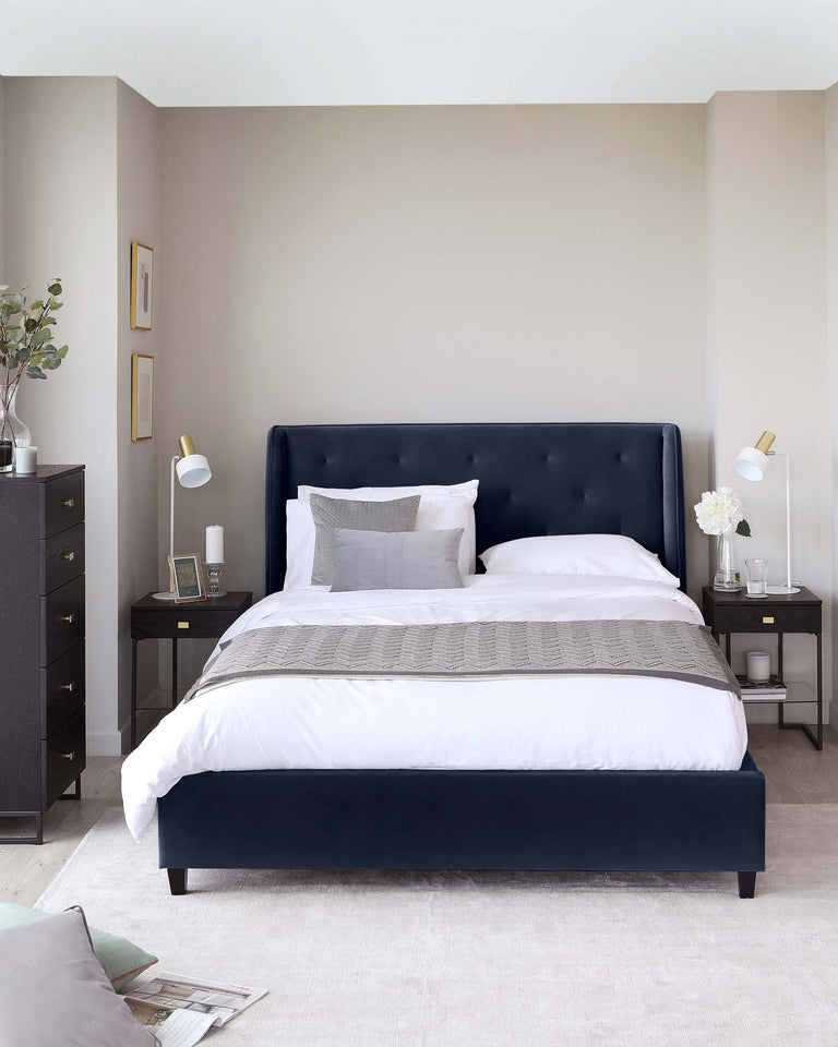 Elegant bedroom interior featuring a navy blue upholstered bed with a tufted headboard, flanked by two matching black wooden nightstands with brass accents. Each nightstand is equipped with a white lamp. There is a grey-toned five-drawer dresser on the left side. The bedding includes crisp white linens accented by a grey textured throw at the foot of the bed. The room is completed by a soft beige carpet and a neutral wall colour, enhancing the sophisticated colour palette.