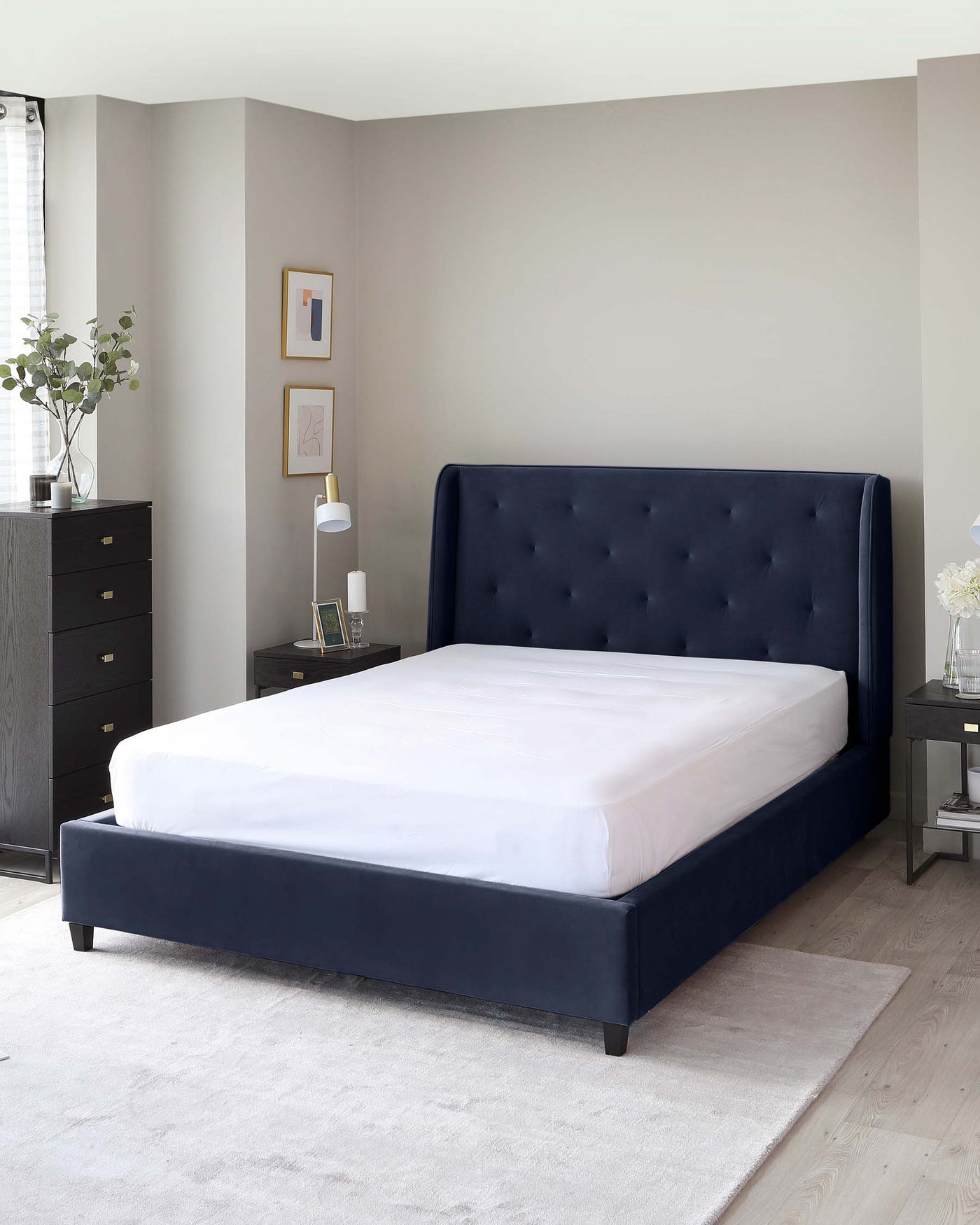 Elegant bedroom furniture set featuring a dark blue upholstered bed with a tufted headboard and matching navy blue bed frame. Beside the bed, a slender nightstand with a white tabletop lamp and a small potted plant. To the left, a taller black chest of drawers with sleek handles complements the ensemble, all displayed on a light grey area rug over wooden flooring.
