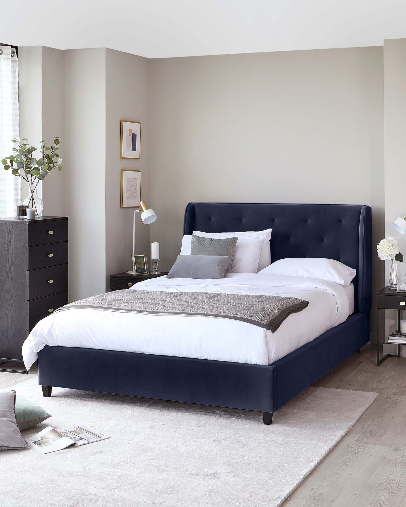 Elegant bedroom scene featuring a navy blue upholstered bed with a tufted headboard, flanked by two matching nightstands with subtle metallic knobs, and a coordinating tall dresser with multiple drawers, all resting on a plush, light grey area rug.