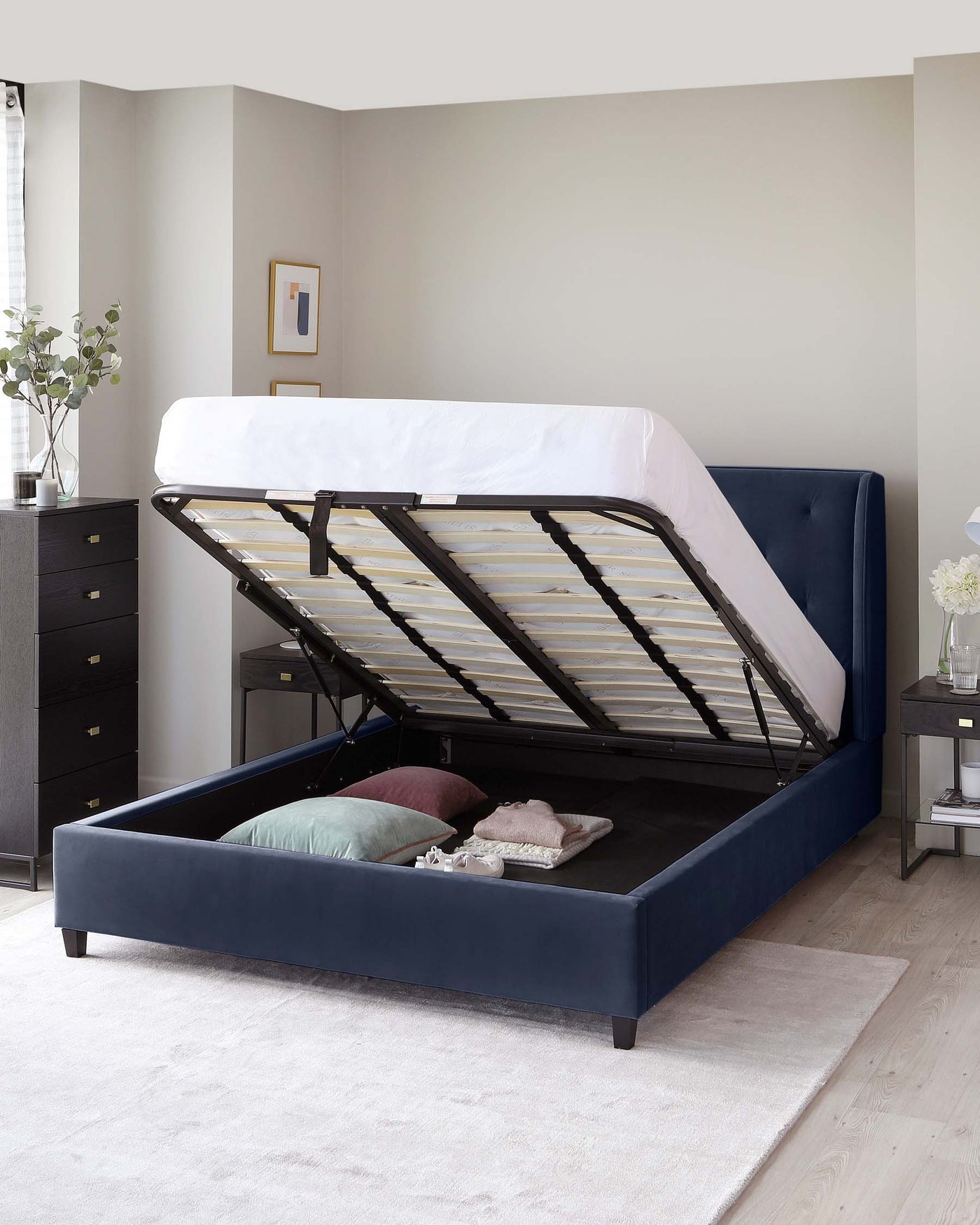 A contemporary upholstered storage bed with a lifted mattress base revealing a spacious storage compartment. The bed features a navy blue finish, a padded headboard, and a bed frame with wooden slats. To the side is a matching dark wood nightstand with drawers. The room is accessorized with a neutral-toned rug, plants, and minimalistic decor.