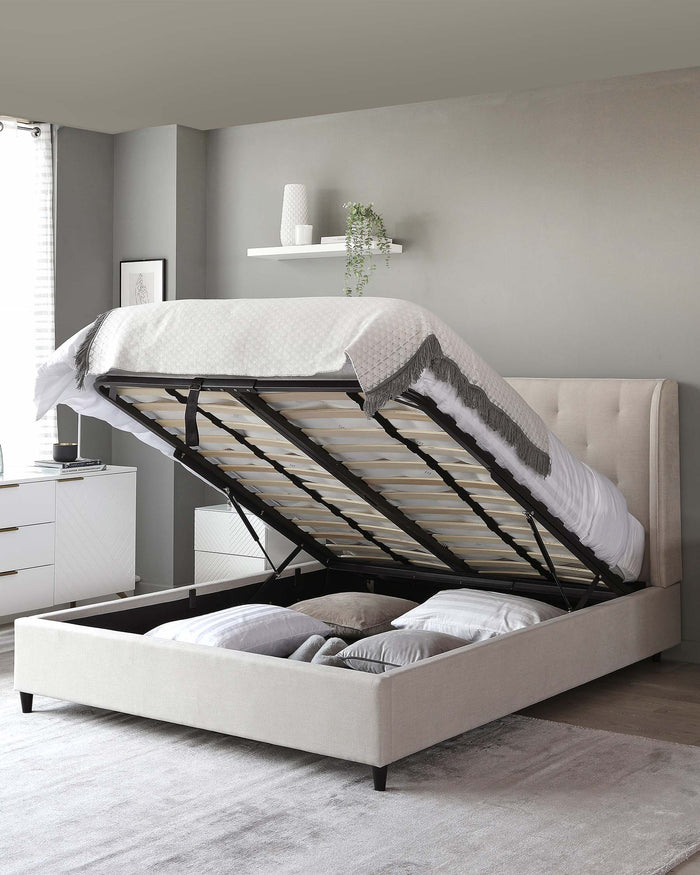 A contemporary beige upholstered storage bed with a lifted mattress base revealing ample under-bed storage, featuring a patterned white bedspread and pillows, set within a modern bedroom with matching beige and white decor.
