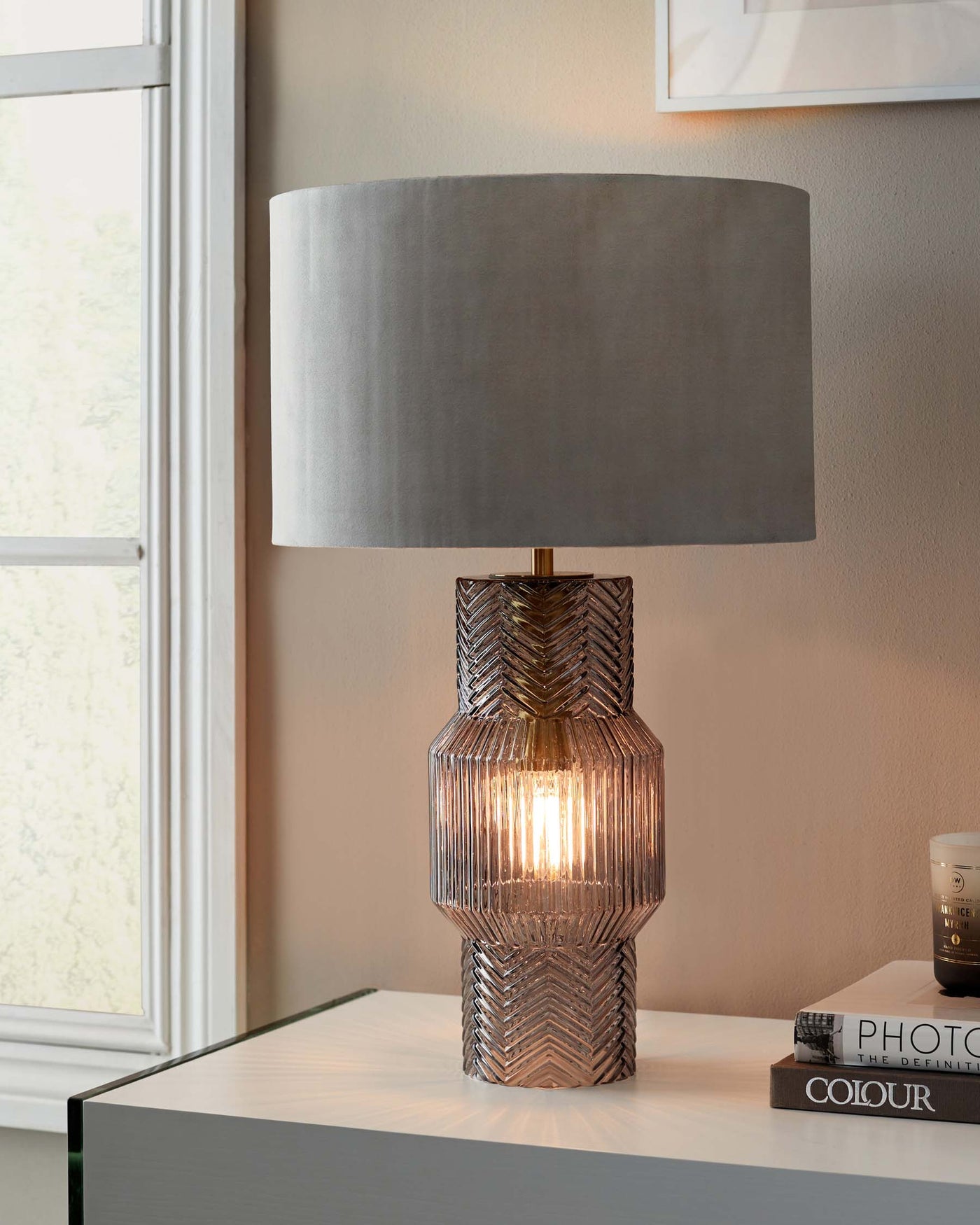 Elegant table lamp with a textured metallic body and a grey cylindrical fabric shade, displayed on a minimalist high-gloss white side table, evoking a modern aesthetic.