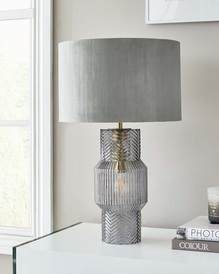 Elegant table lamp with a textured, tiered base featuring a combination of herringbone and vertical ridges, and a transitional brass accent. Topped with a round, grey lampshade that diffuses soft light. Set upon a glossy white surface, possibly a sleek modern side or console table.