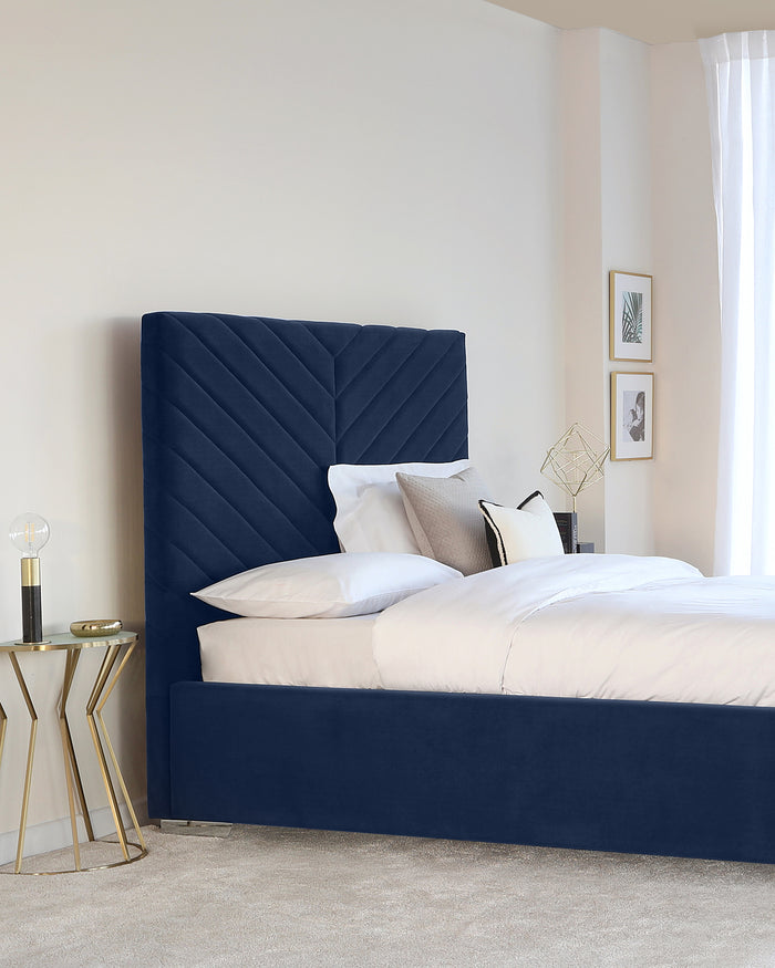 Elegant navy blue upholstered bed with a tall, channel-tufted headboard and matching bed frame, paired with a geometric brass and glass side table holding a modern glass table lamp beside the bed.