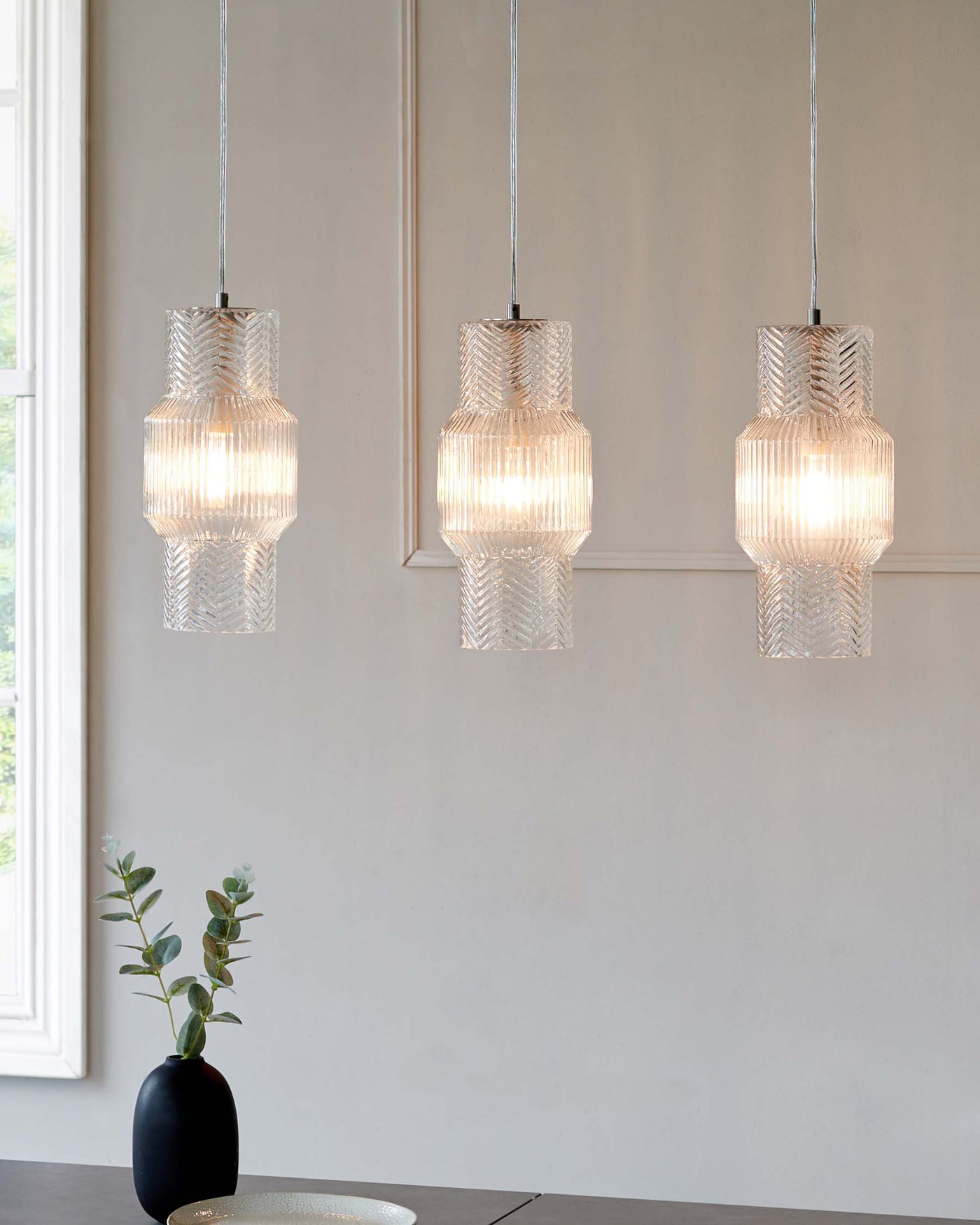 Three textured glass pendant lights hanging in a line above a minimalist dining table with a small black vase and green branches as the centrepiece.