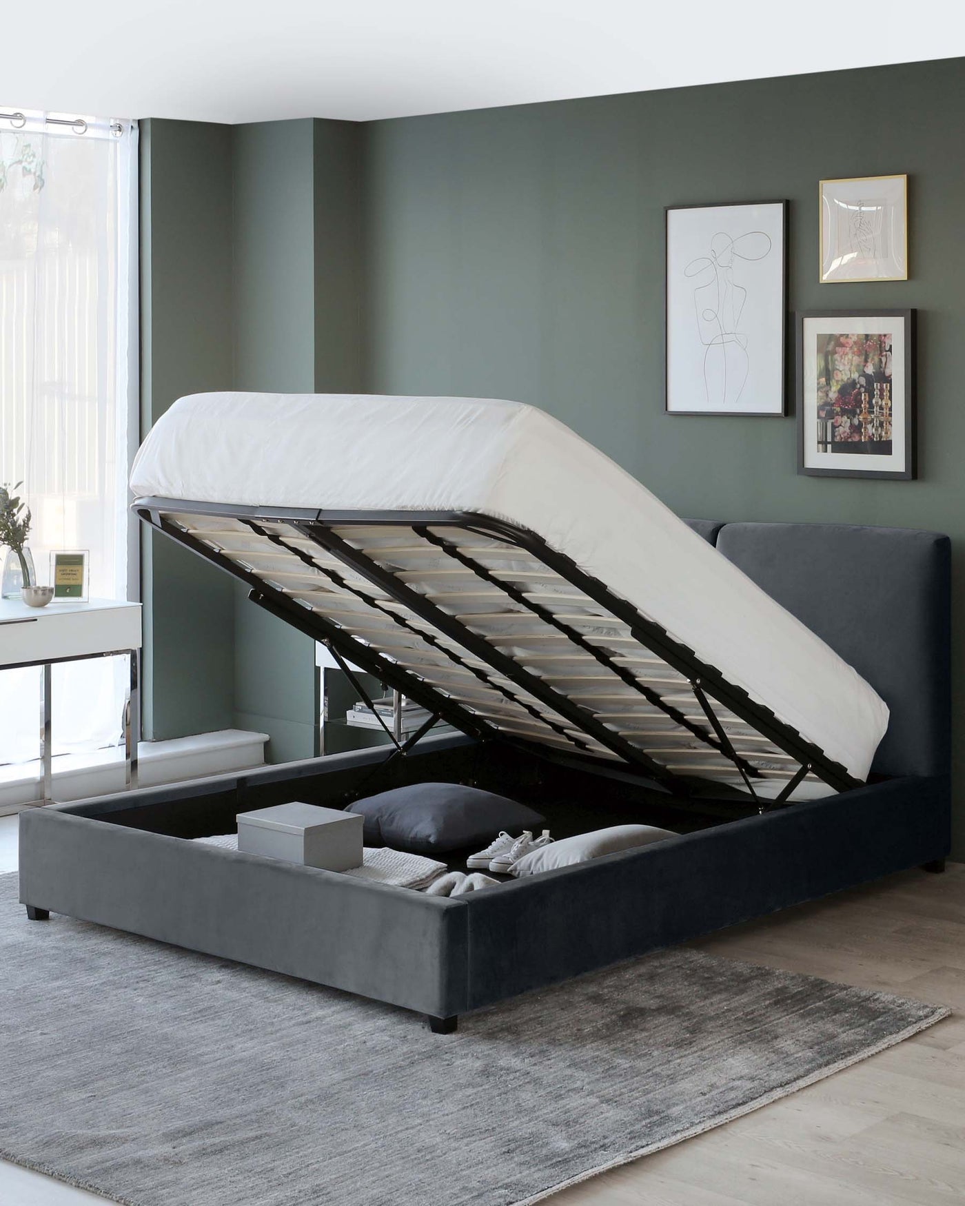 A modern dark grey upholstered storage bed with a lifted mattress base revealing a spacious under-bed storage compartment. The bed features a simple, sleek design and is positioned on a light grey area rug that complements the neutral-toned flooring.
