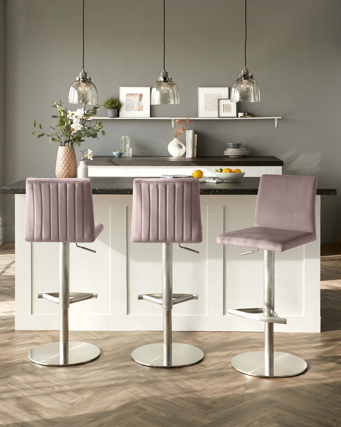 Modern kitchen interior featuring three plush, mauve-pink bar stools with backrests, mounted on sleek, adjustable-height chrome pedestals with circular bases and footrests. The bar stools are placed in front of a kitchen island with a white cabinet and a dark countertop, under elegant glass pendant lights. The shelves above the island display minimalist decorative items.