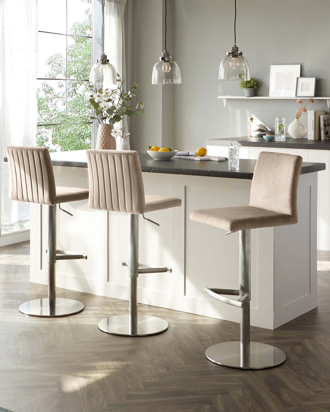 Modern adjustable bar stools with beige upholstered seats, vertical stitching on the backrest, and shiny chrome bases, placed by a kitchen island with a dark countertop.
