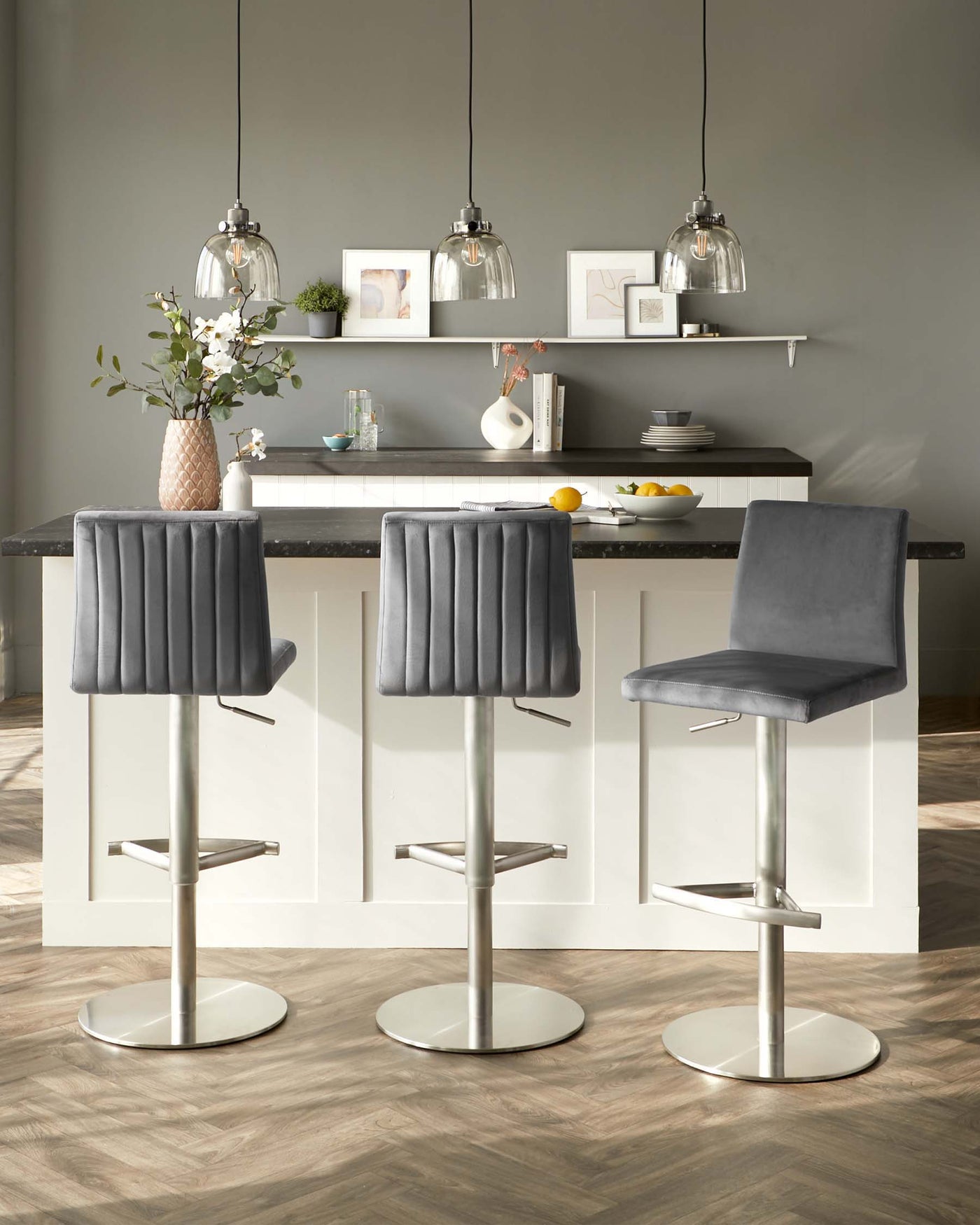 Three modern bar stools with sleek chrome pedestal bases, featuring adjustable height levers and built-in footrests. The seats are upholstered in a luxurious grey fabric with vertical stitching and plush cushioning, offering a comfortable and stylish seating option for a contemporary kitchen island.