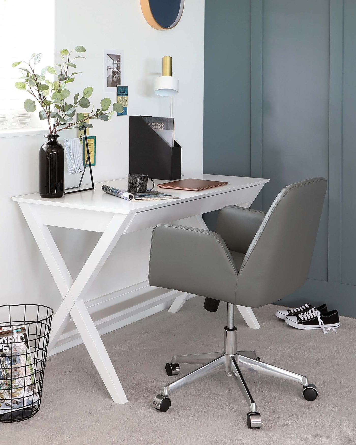 A modern home office setup featuring a sleek, white crisscross-legged desk paired with a sophisticated grey upholstered swivel chair with a high backrest and stainless steel base on casters.