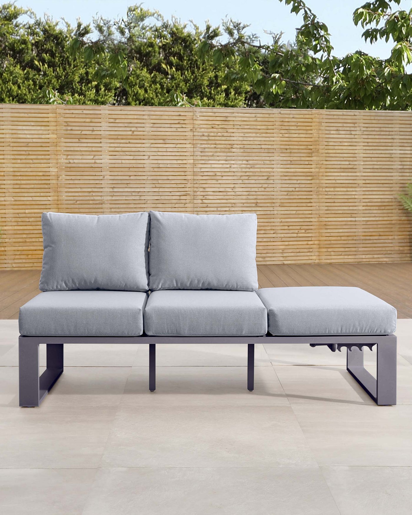 Modern outdoor sectional sofa with grey upholstery and a matching chaise lounge, featuring a sleek dark metal frame, placed on a patio with a bamboo fence background.