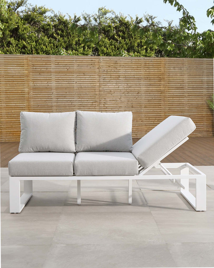 Modern outdoor furniture set comprising a white, minimalist two-seat sofa with grey cushions, paired with a matching white sun lounger with an adjustable backrest and a grey cushion, all staged on a smooth patio surface with a bamboo fence and greenery in the background.