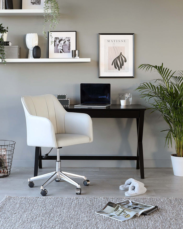 Modern home office setup featuring a sleek black writing desk with a slim silhouette and a minimalist white office chair with padded seat and backrest on a shiny, chrome swivel base with caster wheels. A white floating shelf is displayed above the desk, accented with decorative items and framed pictures. A shaggy area rug lies beneath the chair, and a potted indoor plant adds a touch of greenery to the space.