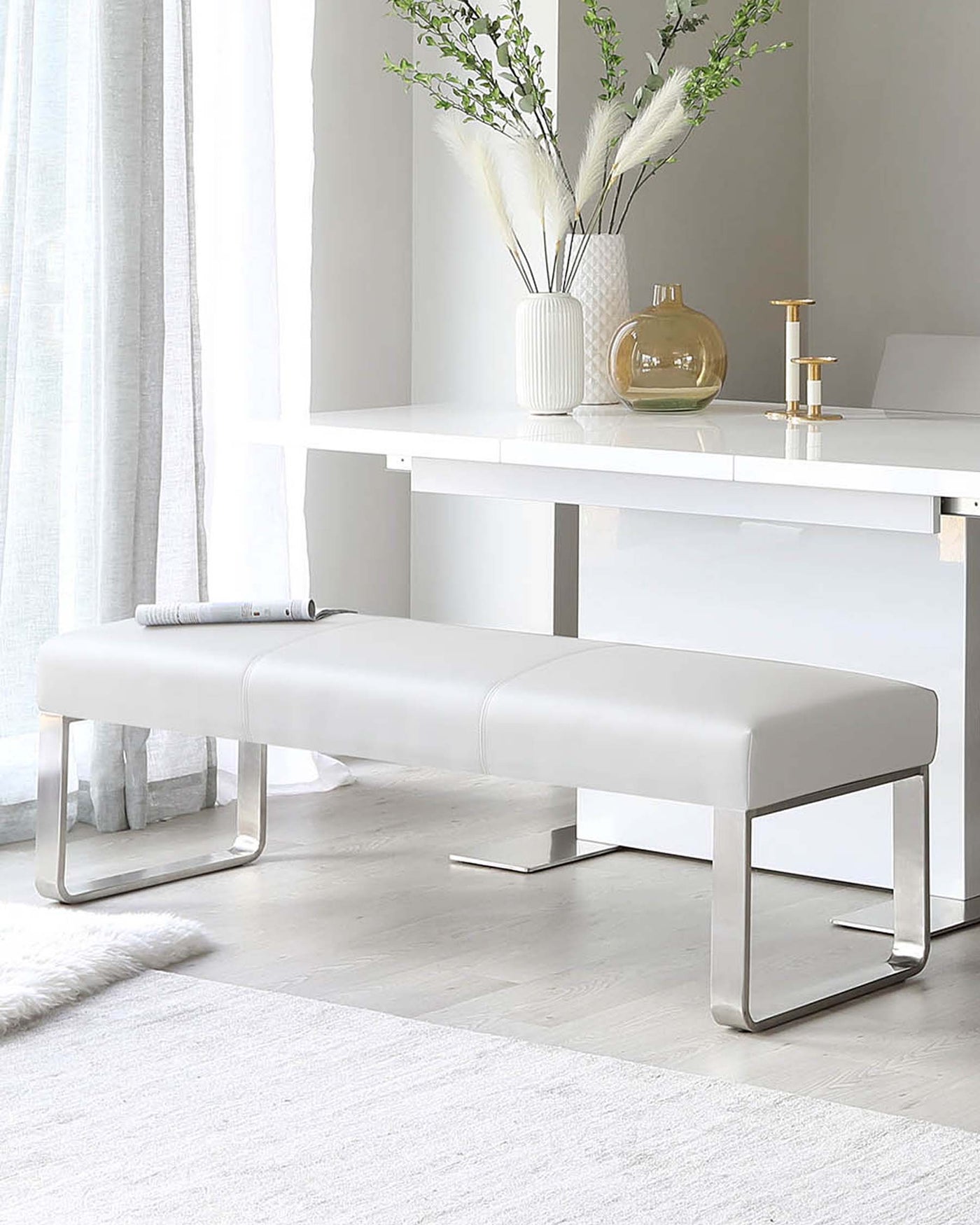 Modern minimalist furniture set including a sleek white rectangular console table with metal supports and a matching bench with a padded leather seat and metallic base. The scene is complemented by decorative vases and a small gold candlestick holder on the table, indicating a contemporary and elegant design aesthetic.