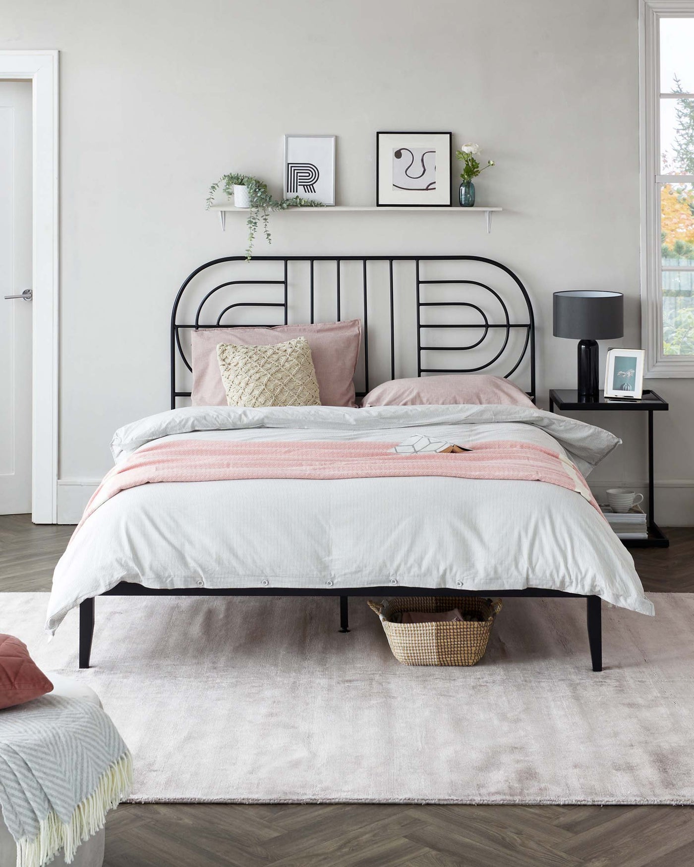 A contemporary-style bedroom featuring a metal frame bed with a minimalist headboard design, complemented by soft pink and grey bedding. Next to the bed stands a sleek black bedside table with a modern lamp, all anchored upon a plush, light grey area rug. A small wicker basket can be observed under the bed.