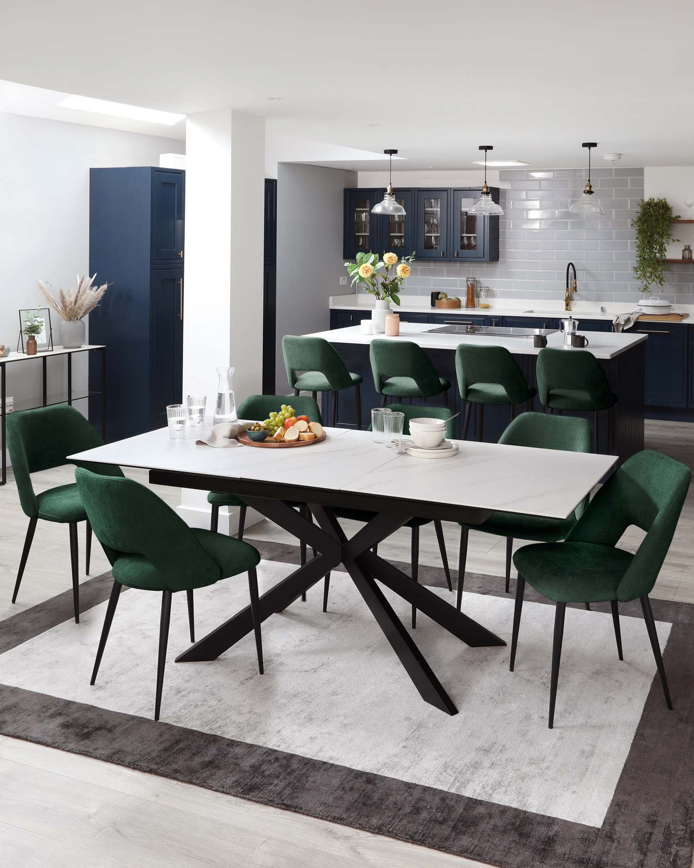 Modern dining room featuring a rectangular white dining table with a unique black geometric base. Surrounded by six plush green velvet chairs with black legs. The ensemble sits atop a two-tone grey area rug.
