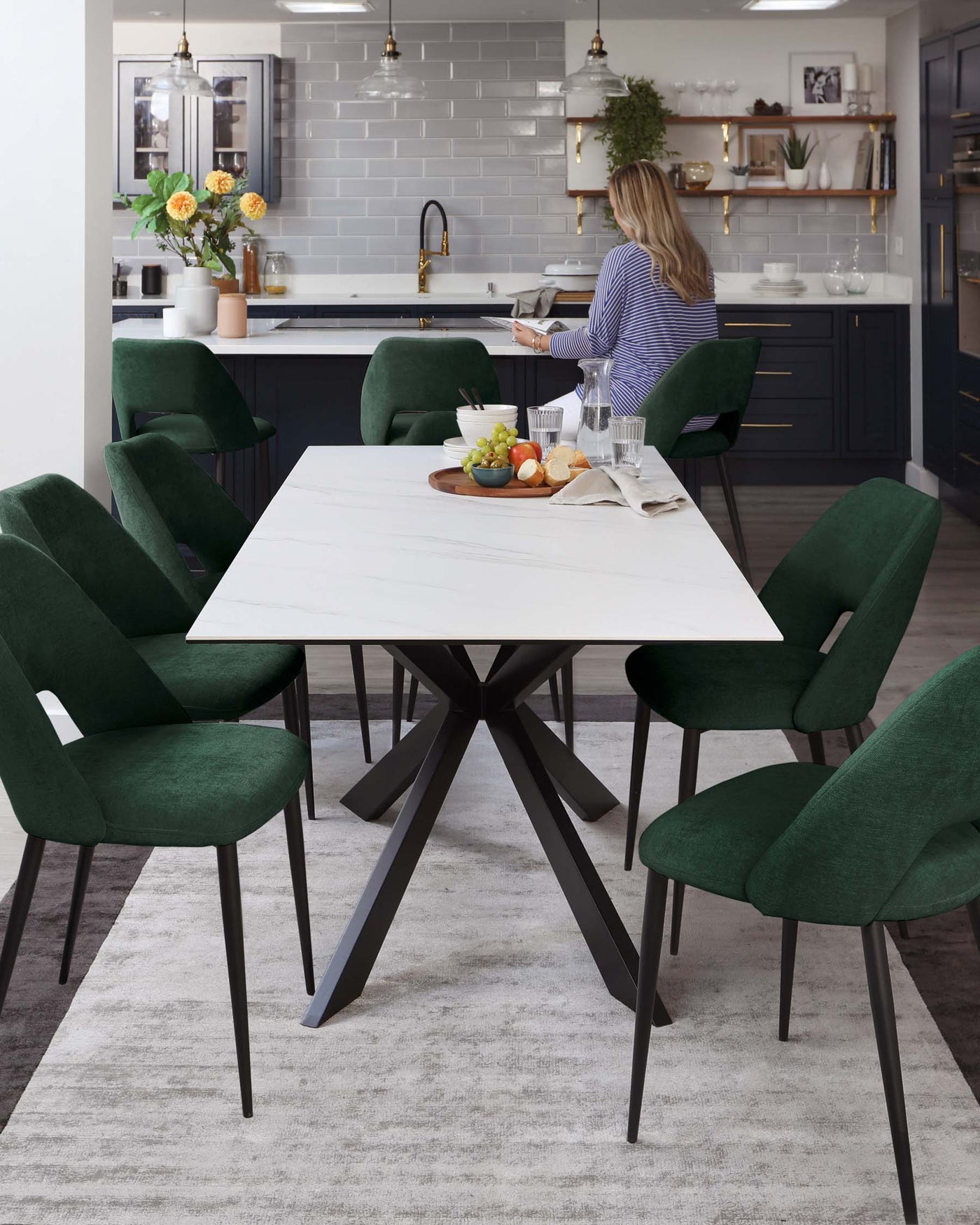 Modern dining room with a white marble-topped table featuring an angular black metal base, surrounded by six plush, forest green velvet dining chairs with sleek black legs, atop a textured grey area rug.