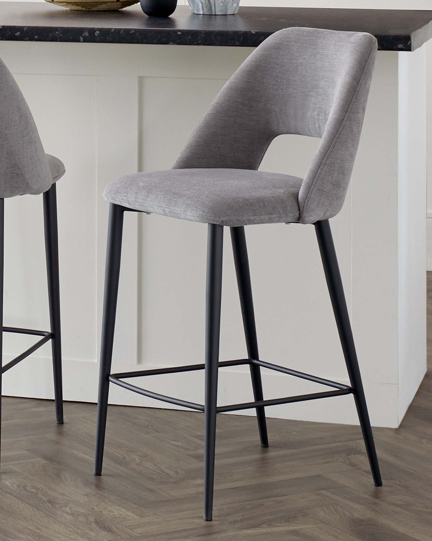 Modern bar stool with a curved back, light grey fabric upholstery, and slender black metal legs with a circular footrest. The seat is positioned against a kitchen island with a contrasting dark countertop.