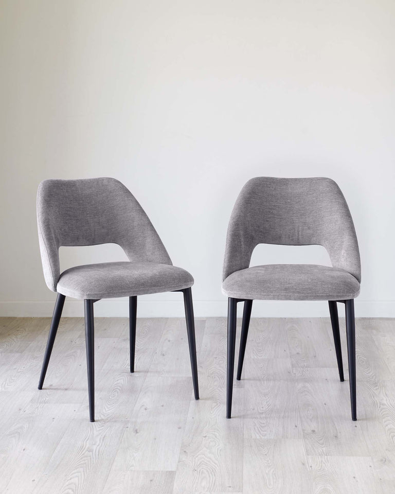 Two modern minimalist dining chairs with grey upholstery and black metal legs on a light wooden floor against a white background.
