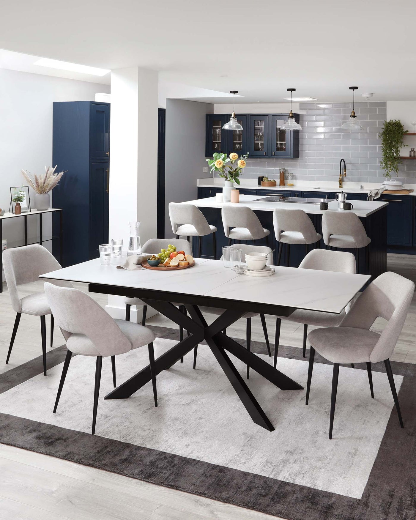 Modern dining room furniture featuring a rectangular marble-top table with an angular black metal base, surrounded by six grey upholstered chairs with black metal legs, all placed on a two-tone grey area rug. A sleek kitchen island can be seen in the background.