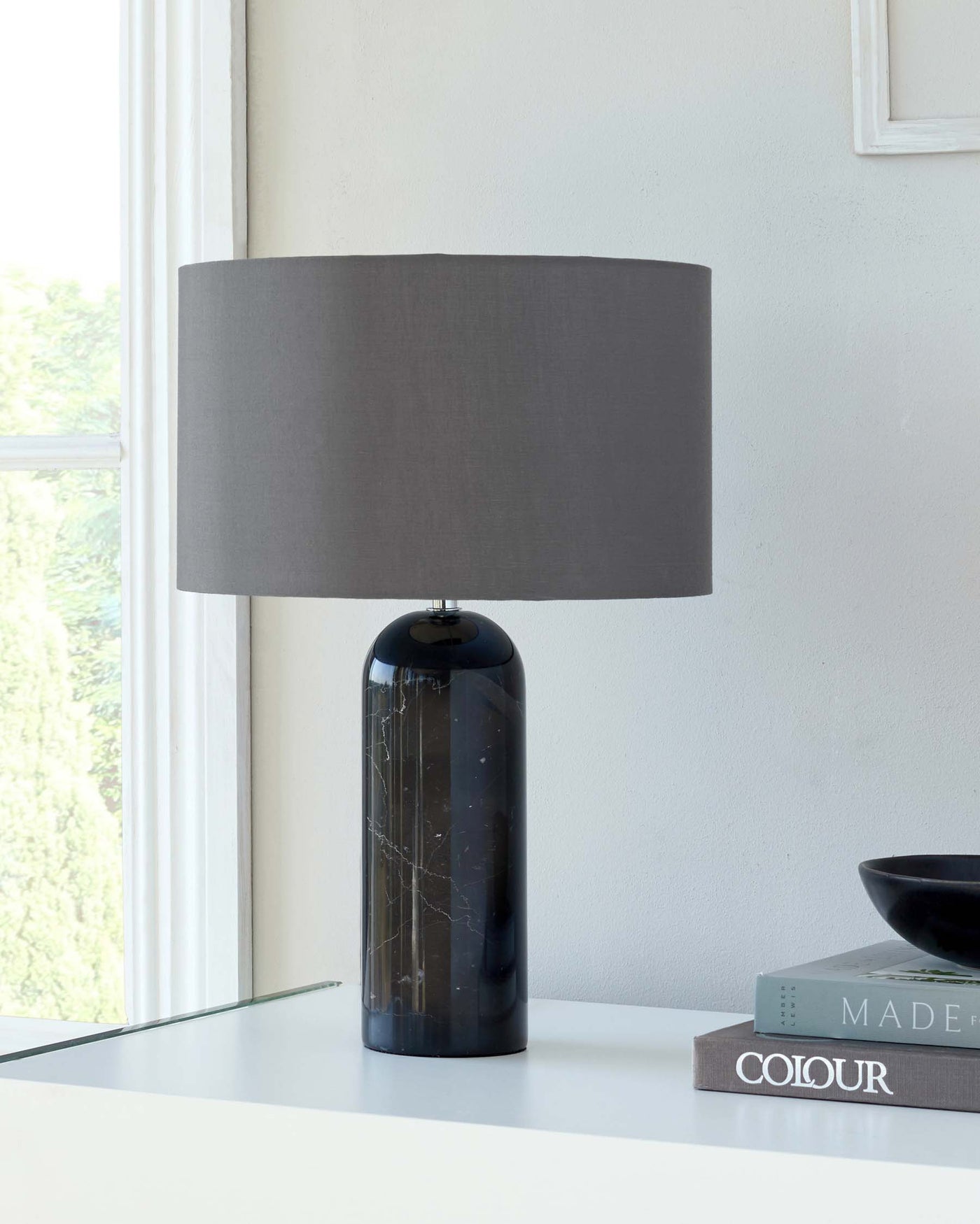 Modern table lamp with a cylindrical, navy blue marble base and a broad, grey fabric drum shade, displayed on a sleek white table next to decorative books and a bowl, with a window providing natural light in the background.