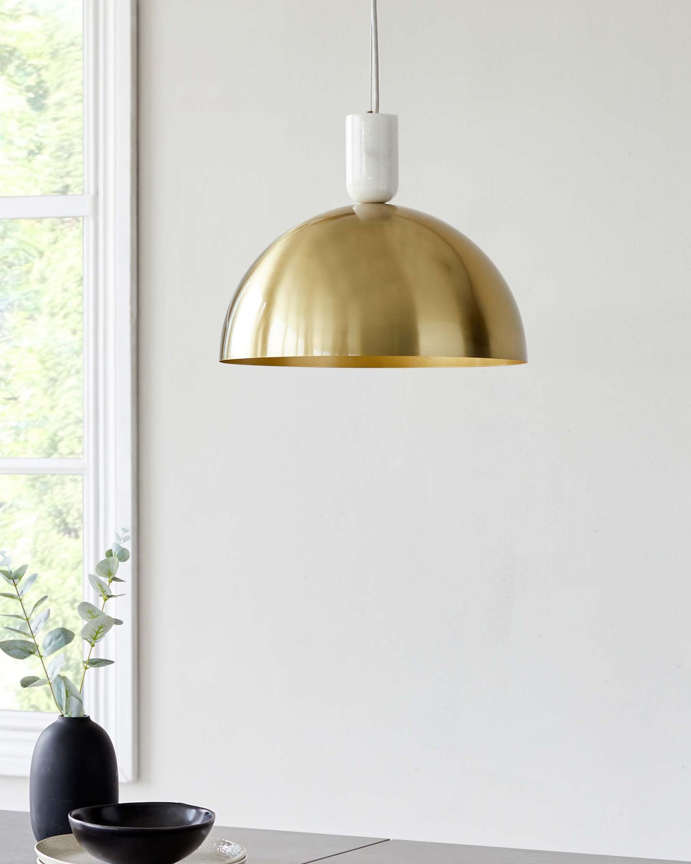 Elegant brass dome pendant light hanging above a minimalist setting including a matte black vase with greenery and a black bowl on a sleek, modern table with a neutral-tone surface.