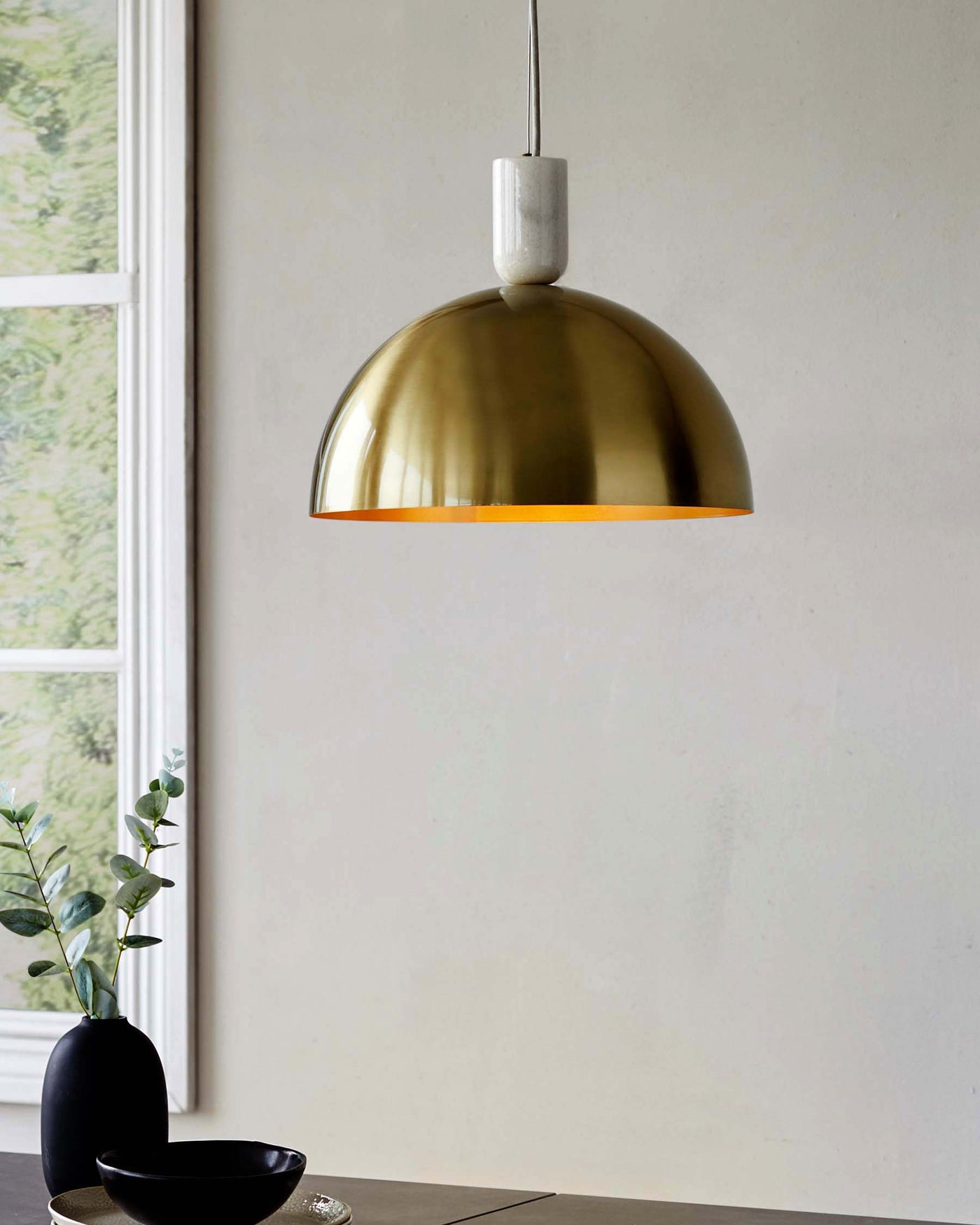 Elegant gold dome pendant light with a matte white top cap, hanging over a sleek dark tabletop adorned with a minimalist black vase containing eucalyptus branches.