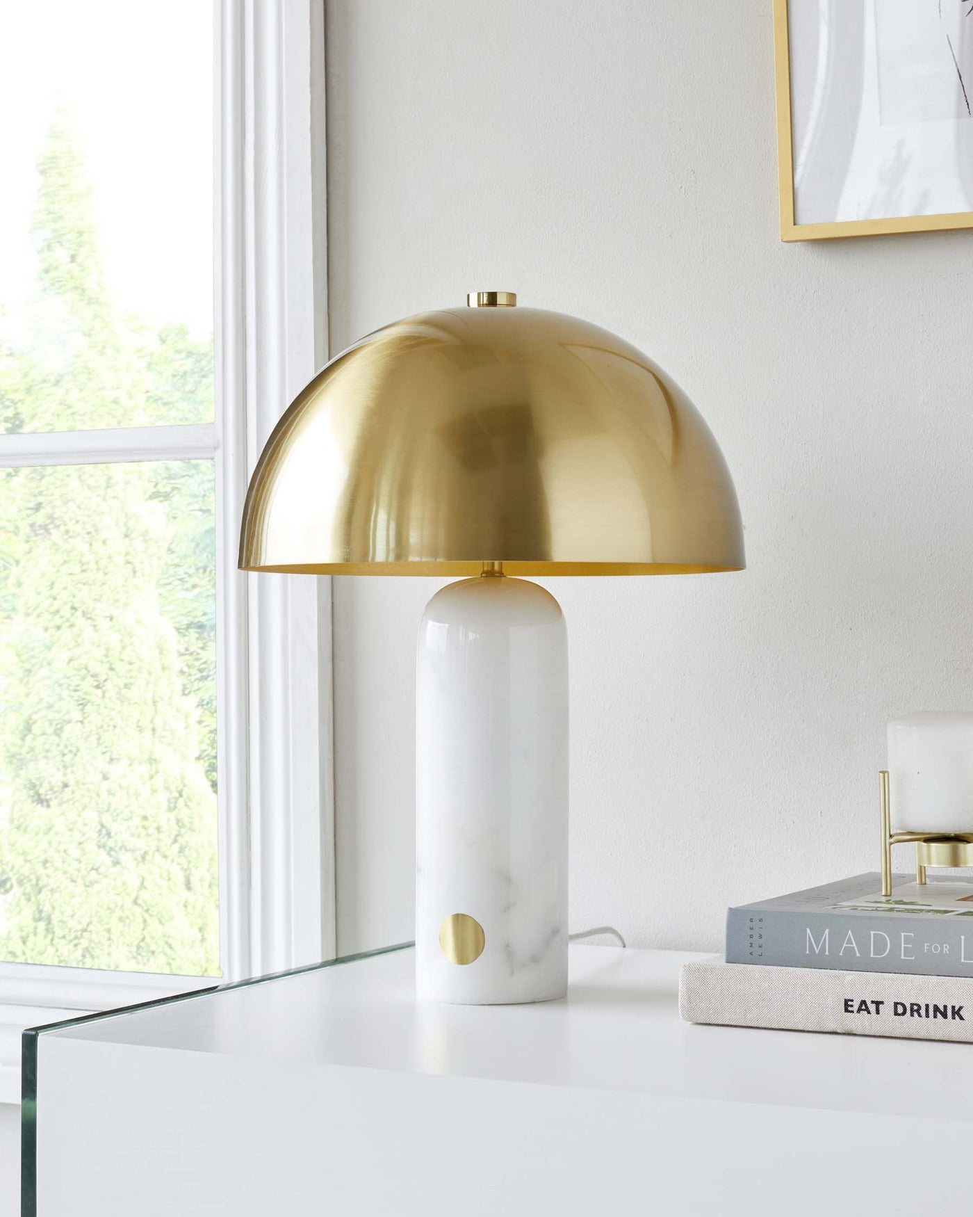 Elegant contemporary table lamp with a glossy brass dome shade and a cylindrical white marble base featuring a gold accent. Positioned on a sleek white tabletop beside a stack of hardcover books.