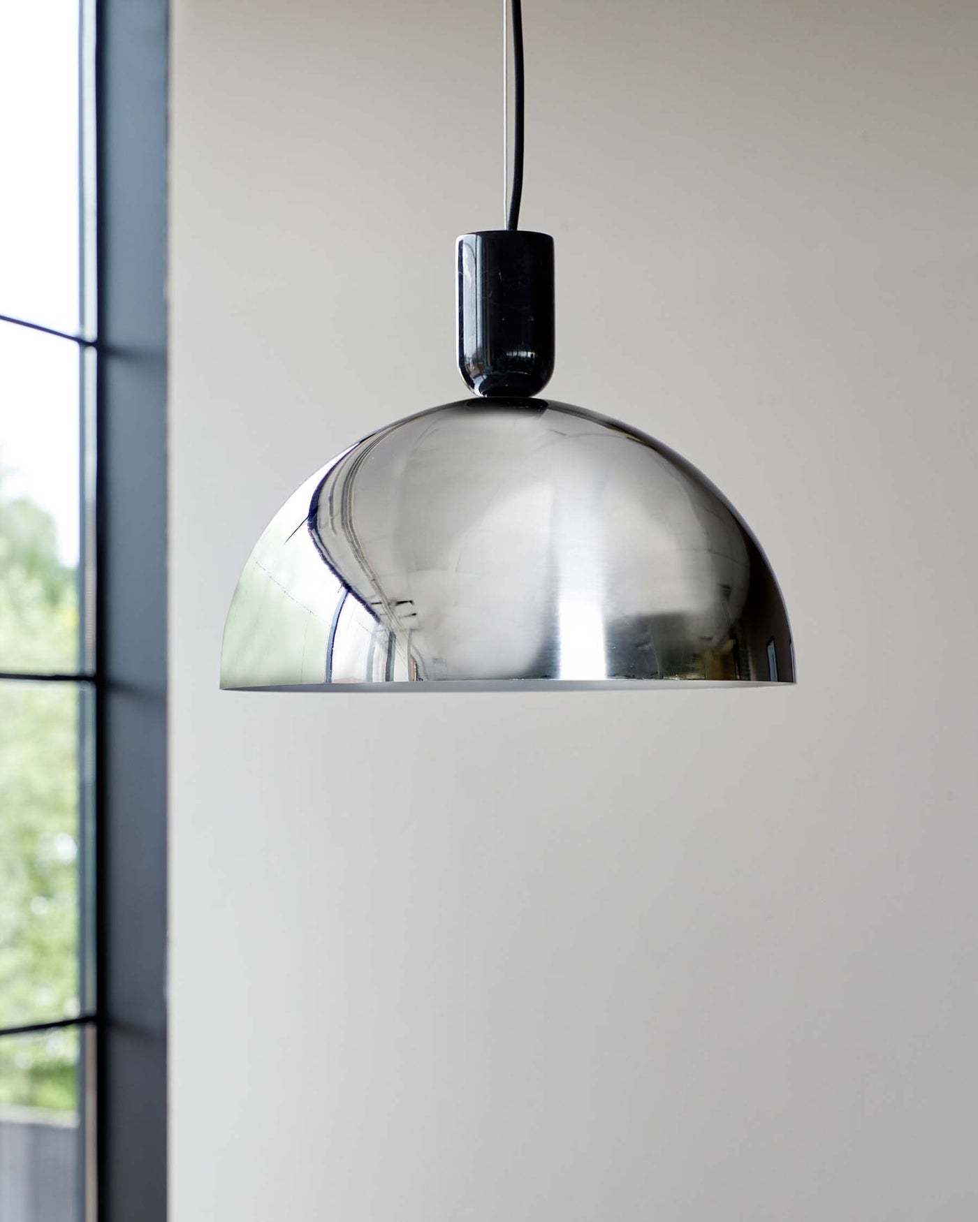 Modern pendant light with a polished chrome dome-shaped shade hanging from a black cord against a neutral background with a windowpane to the left.