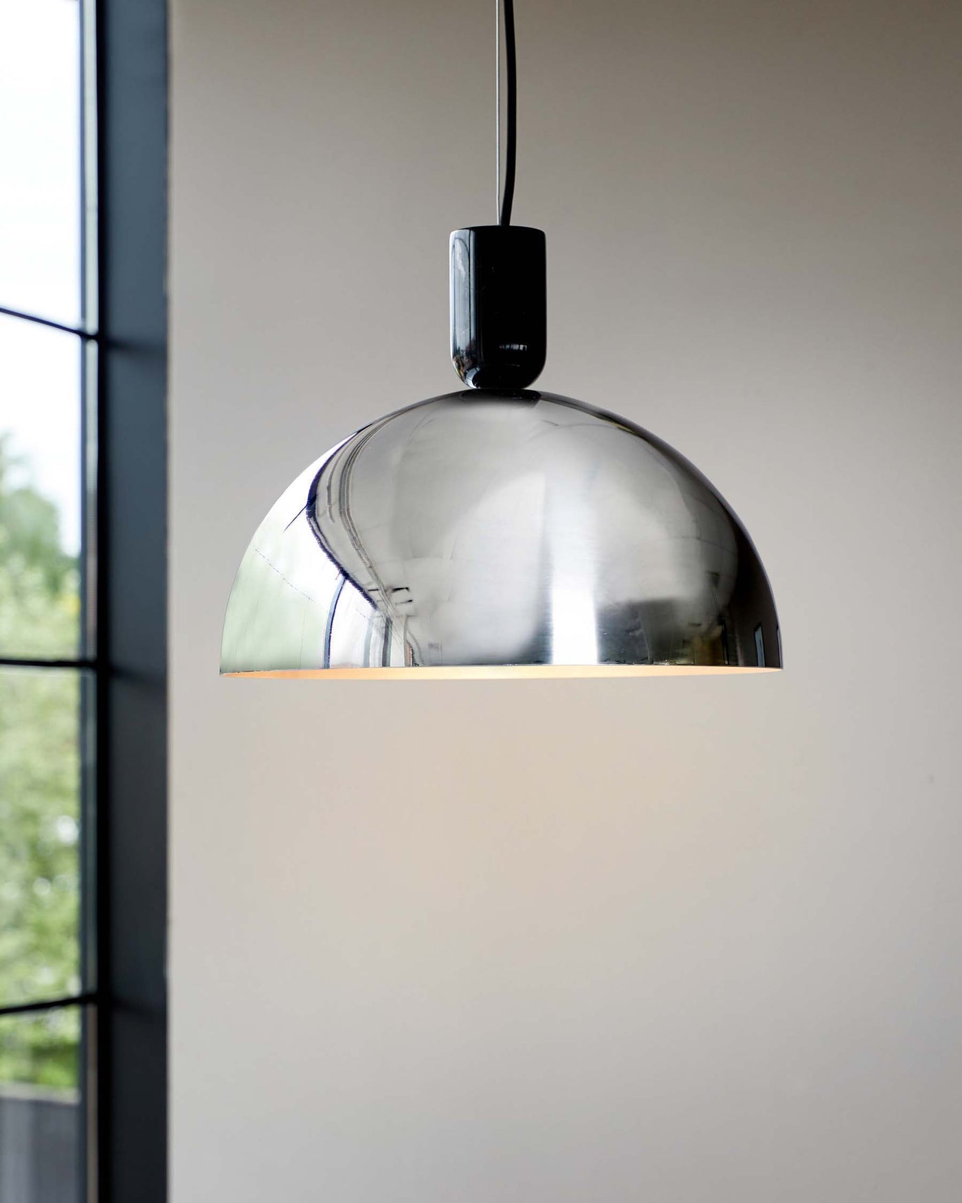 Modern chrome pendant light with a sleek, reflective dome-shaped shade, suspended from a black cable. The fixture is set against a soft neutral background with natural light coming through a window to the side.
