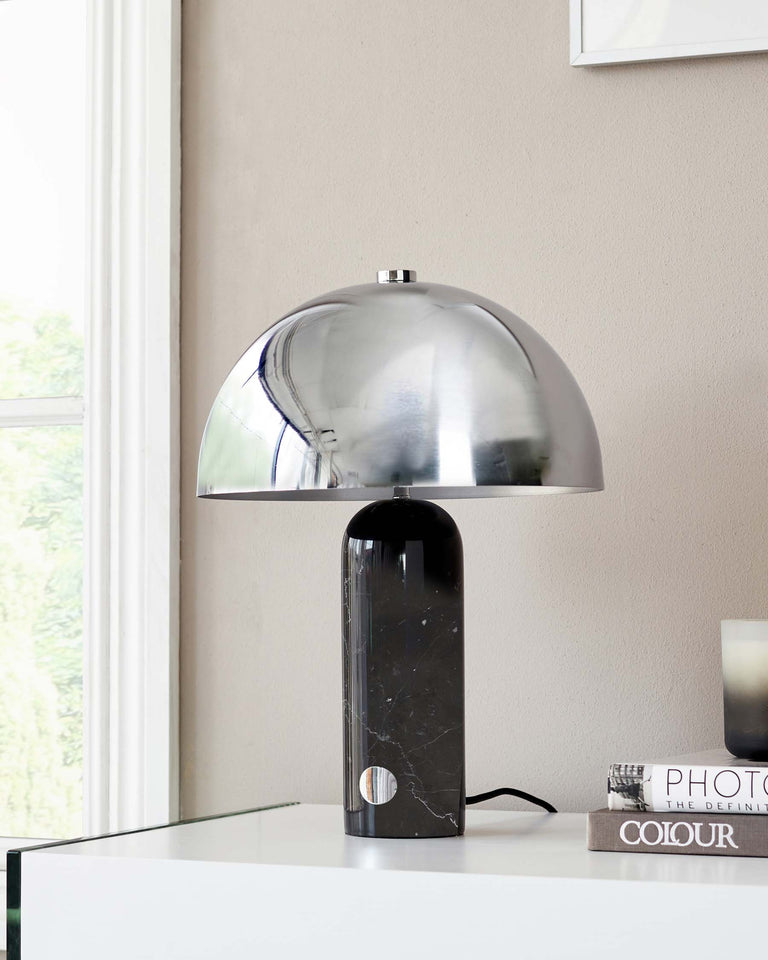 Modern chrome dome table lamp with a polished finish on a marble base, placed on a glossy white table beside stacked books and a framed artwork, with a window providing natural light in the background.