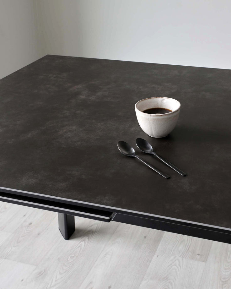 Modern minimalist dark brown dining table with a matte finish and sturdy rectangular legs, complemented by a simple ceramic bowl and a pair of spoons on top.