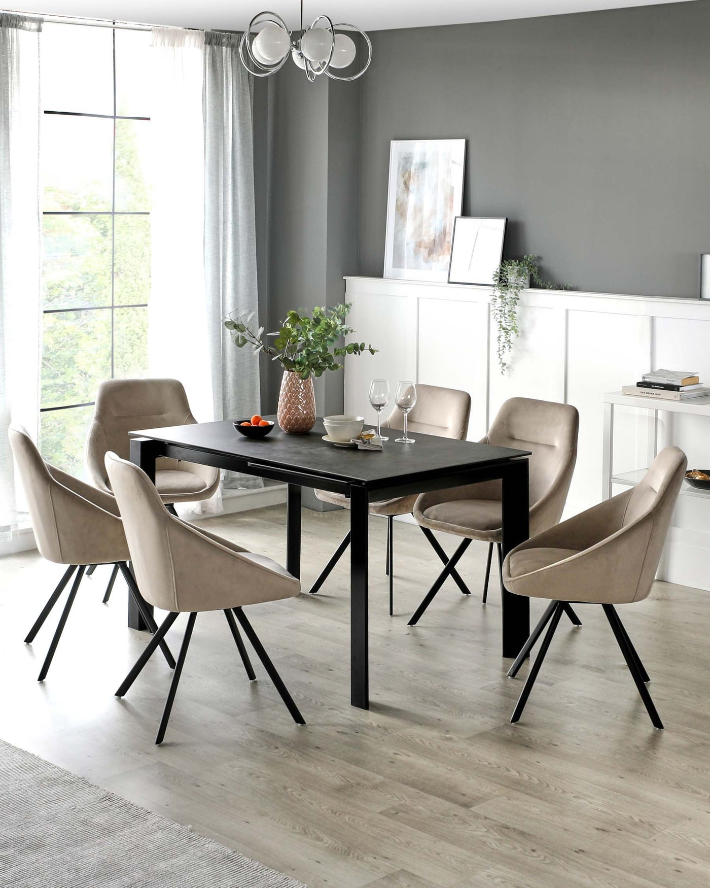 Modern dining room featuring a sleek black rectangular table with a smooth finish, surrounded by six upholstered chairs in a light taupe fabric. The chairs have a curved backrest and are supported by black metal legs that complement the table. A minimalist white sideboard with decorative items sits against the wall.