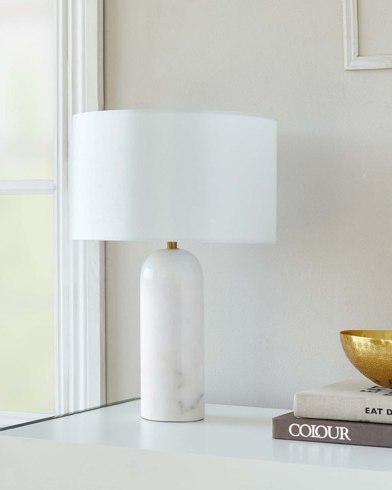 Elegant modern table lamp with a white marble base and a broad cylindrical white shade, displayed on a glossy glass tabletop with decorative books and a gold bowl.