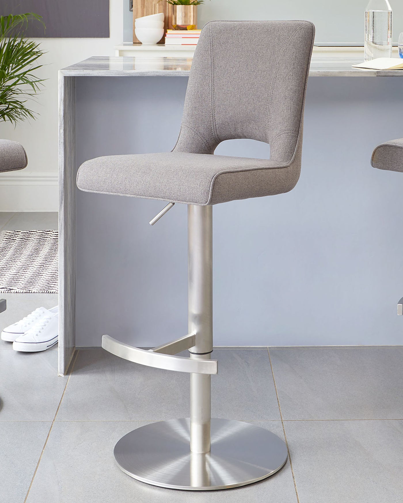 Modern bar stool with a grey upholstered seat and a curved backrest, featuring a sleek metallic pedestal base with a built-in footrest and a round, flat base plate. The stool's height appears to be adjustable, suggested by the visible lever under the seat.