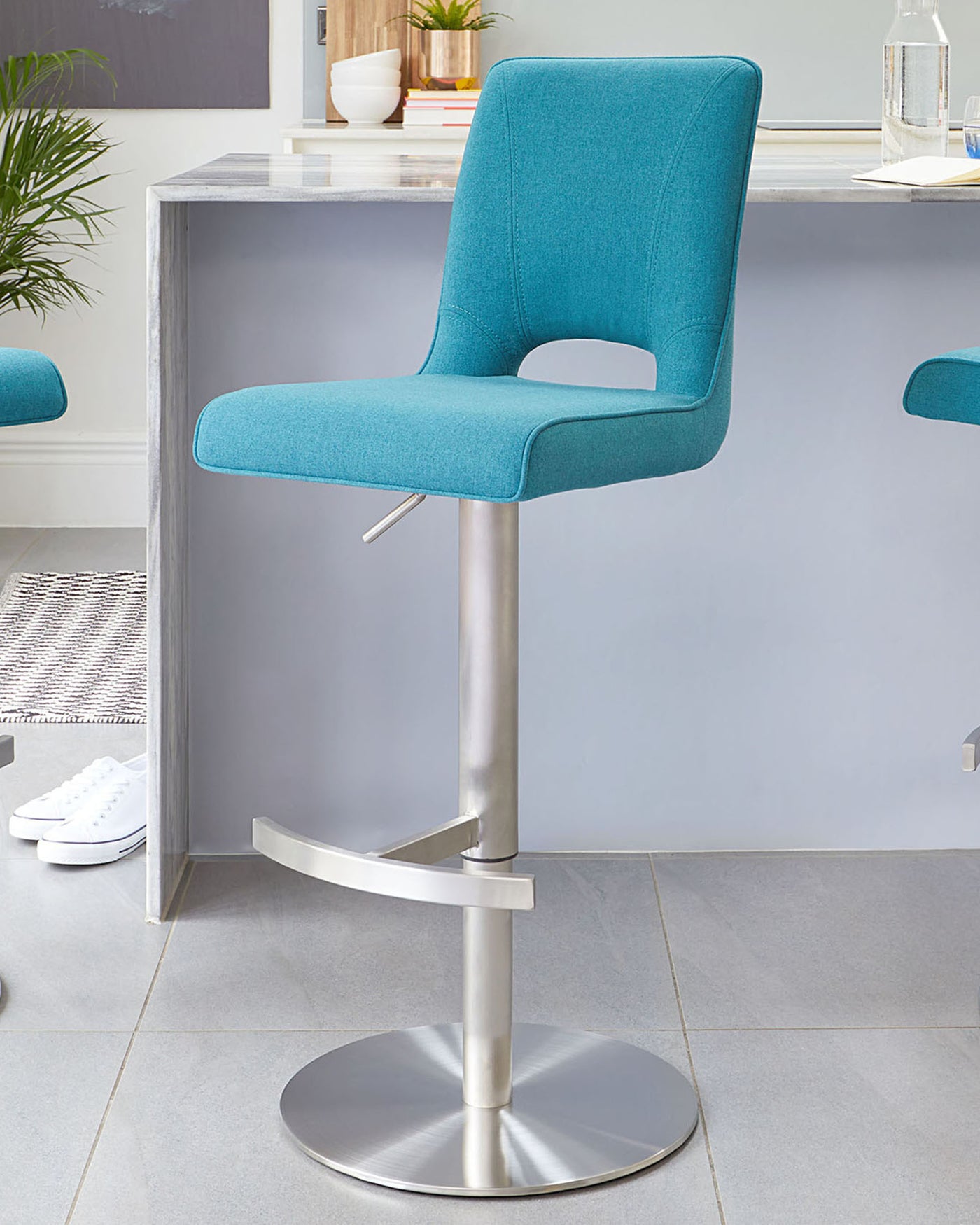 Modern adjustable-height bar stool with a turquoise blue fabric upholstery, featuring an ergonomically curved seat and backrest. The stool has a sleek chrome pedestal base with a built-in footrest and a round flat base for stability.